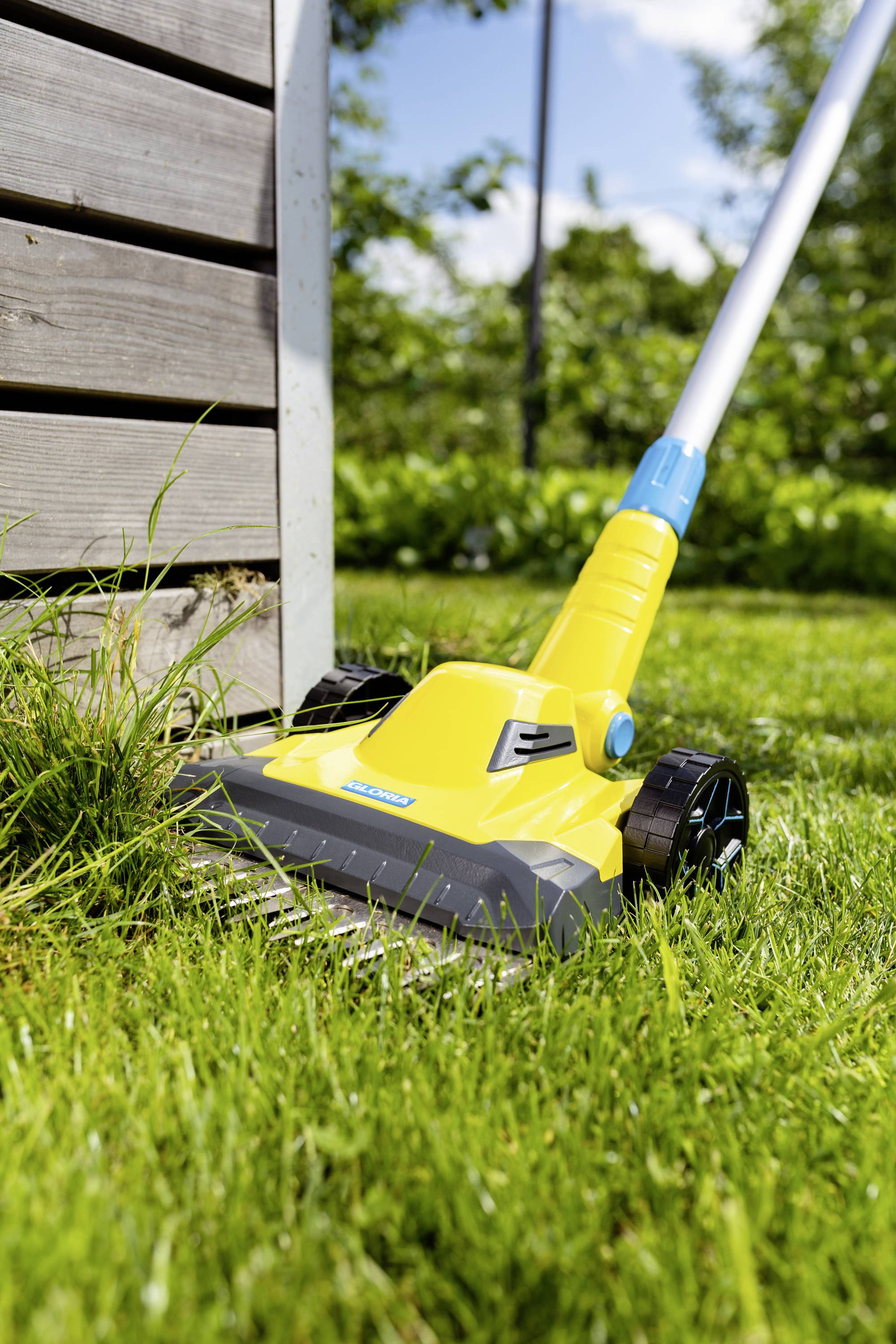 'Ein gelber Rasenkantenschneider trimmt Gras entlang einer Holzwand im sonnigen Garten.'