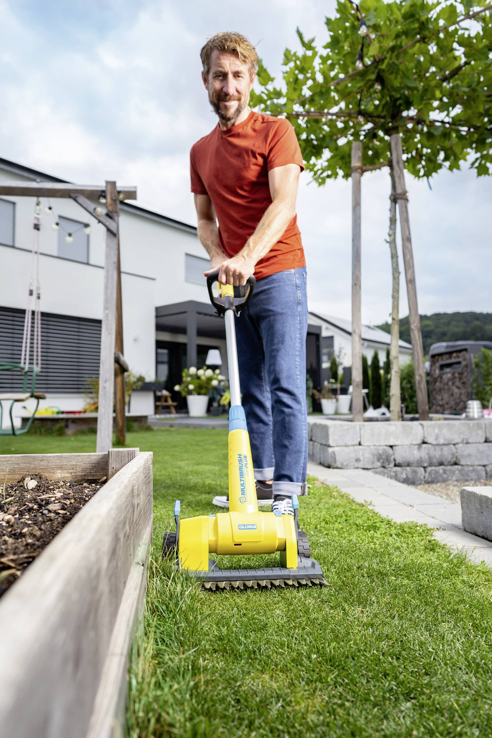 Ein Mann mit rotem Shirt mäht den Rasen in einem Garten neben einem erhöhten Blumenbeet. Im Hintergrund ist ein modernes Haus zu sehen.