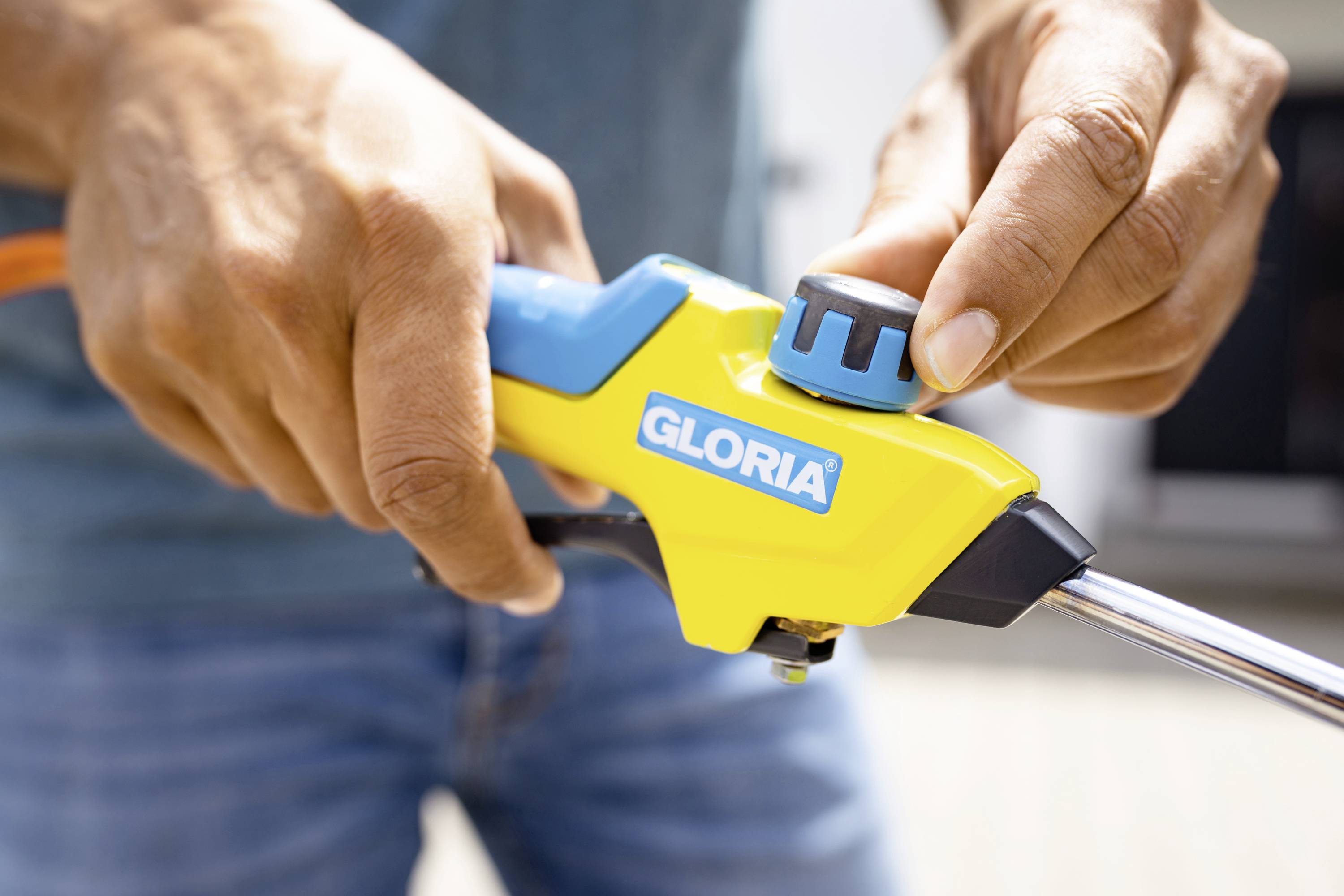 Person bedient ein gelbes Gartenwerkzeug mit blauer Düse und Aufschrift 'Gloria'. Hände drehen am Einstellknopf.