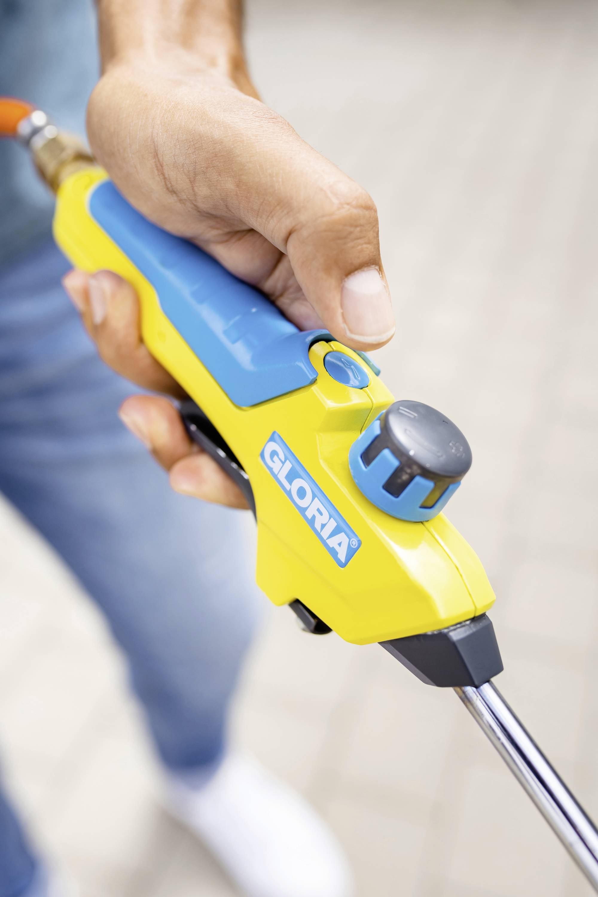 Eine Hand hält ein gelbes Gartengerät mit blauer Beschriftung 'GLORIA'. Es scheint für das Sprühen oder Spritzen von Wasser gedacht zu sein.