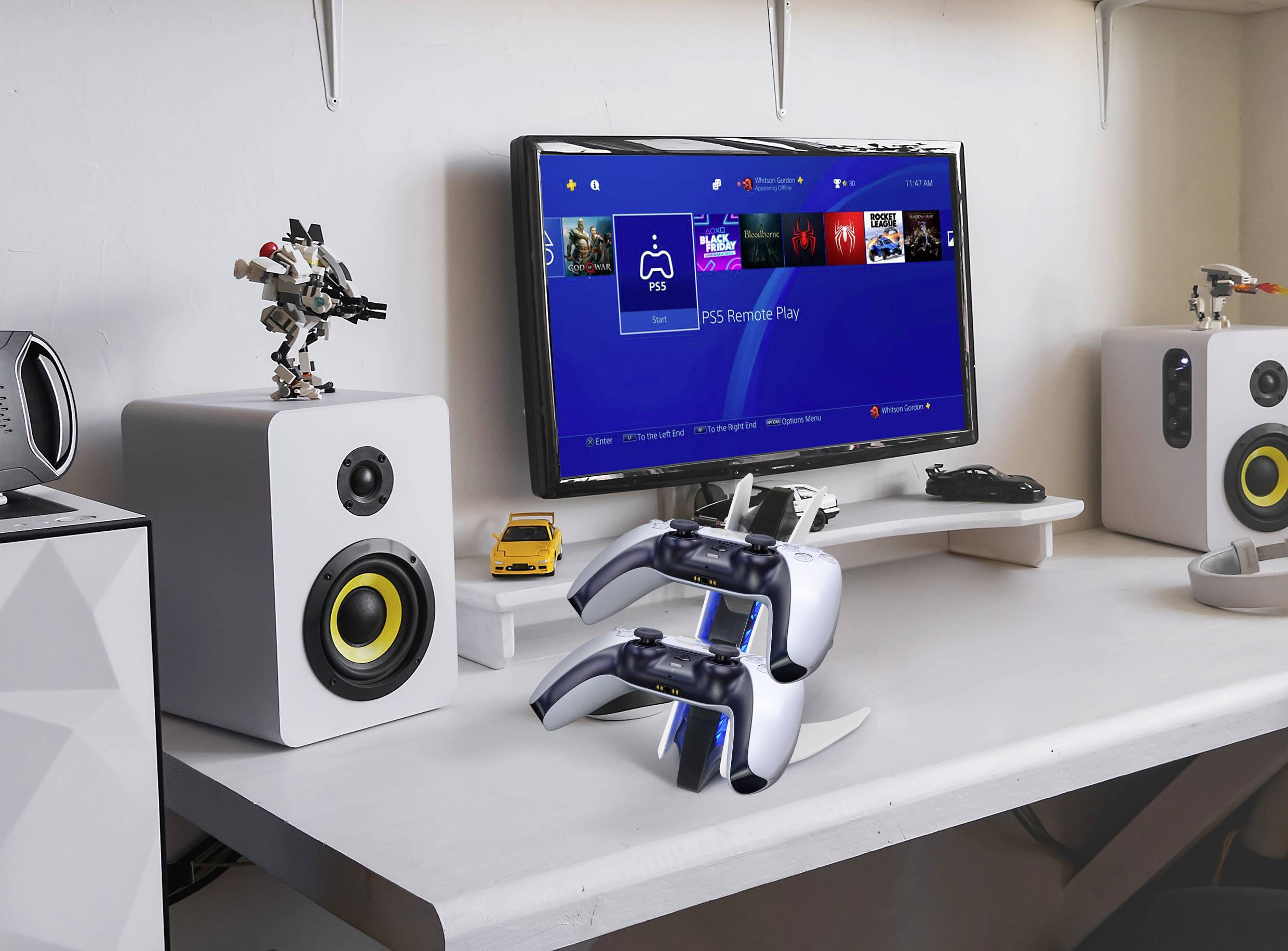 Schreibtisch mit Lautsprechern, einem Monitor mit PS5-Benutzeroberfläche und zwei PS5-Controllern auf einem Ladeständer.
