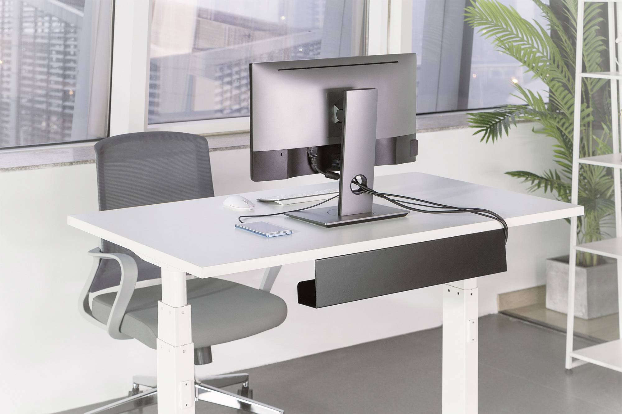 Ein moderner Schreibtisch mit einem Computerbildschirm, Tastatur, und Smartphone. Im Hintergrund sind Fenster mit Stadtblick.