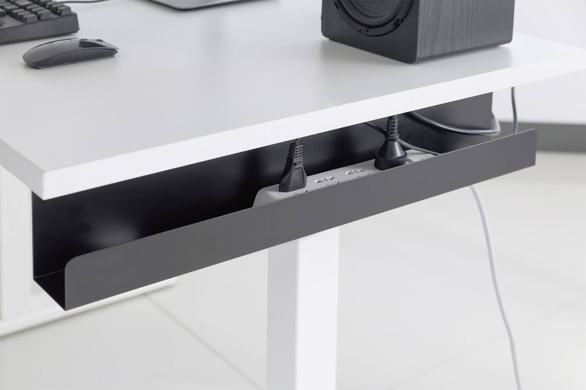 Ein Schreibtisch mit einem Kabelmanagement-Fach voller Kabel und Steckdosenleisten darunter. Oben auf dem Tisch steht ein Lautsprecher.