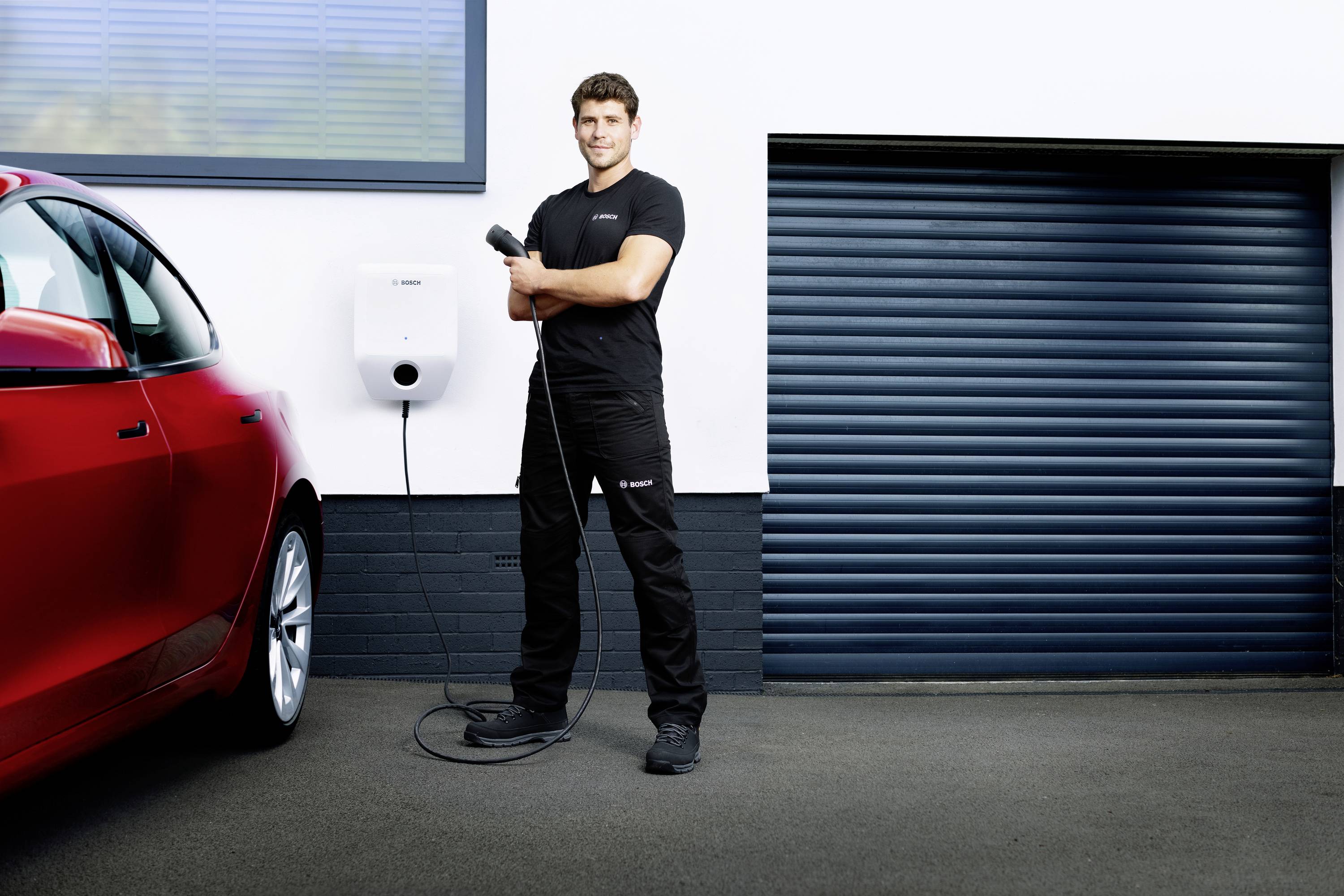 Ein Mann steht neben einem roten Auto und hält ein Ladekabel vor einer blauen Garagentür mit einer an der Wand montierten Ladestation.