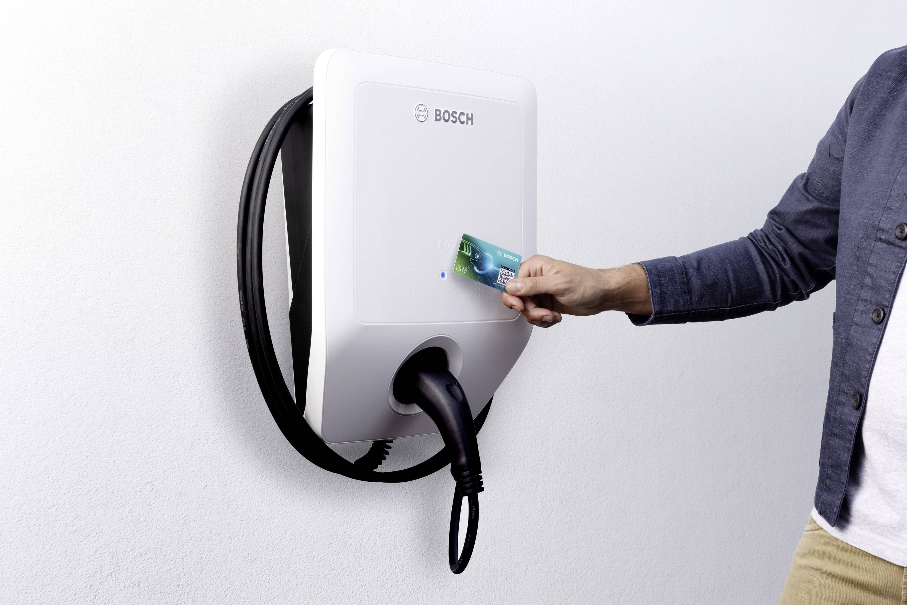 Person hält eine Karte vor ein weißes Bosch-Ladegerät an einer Wand. Ein Kabel ist am Gerät aufgerollt.