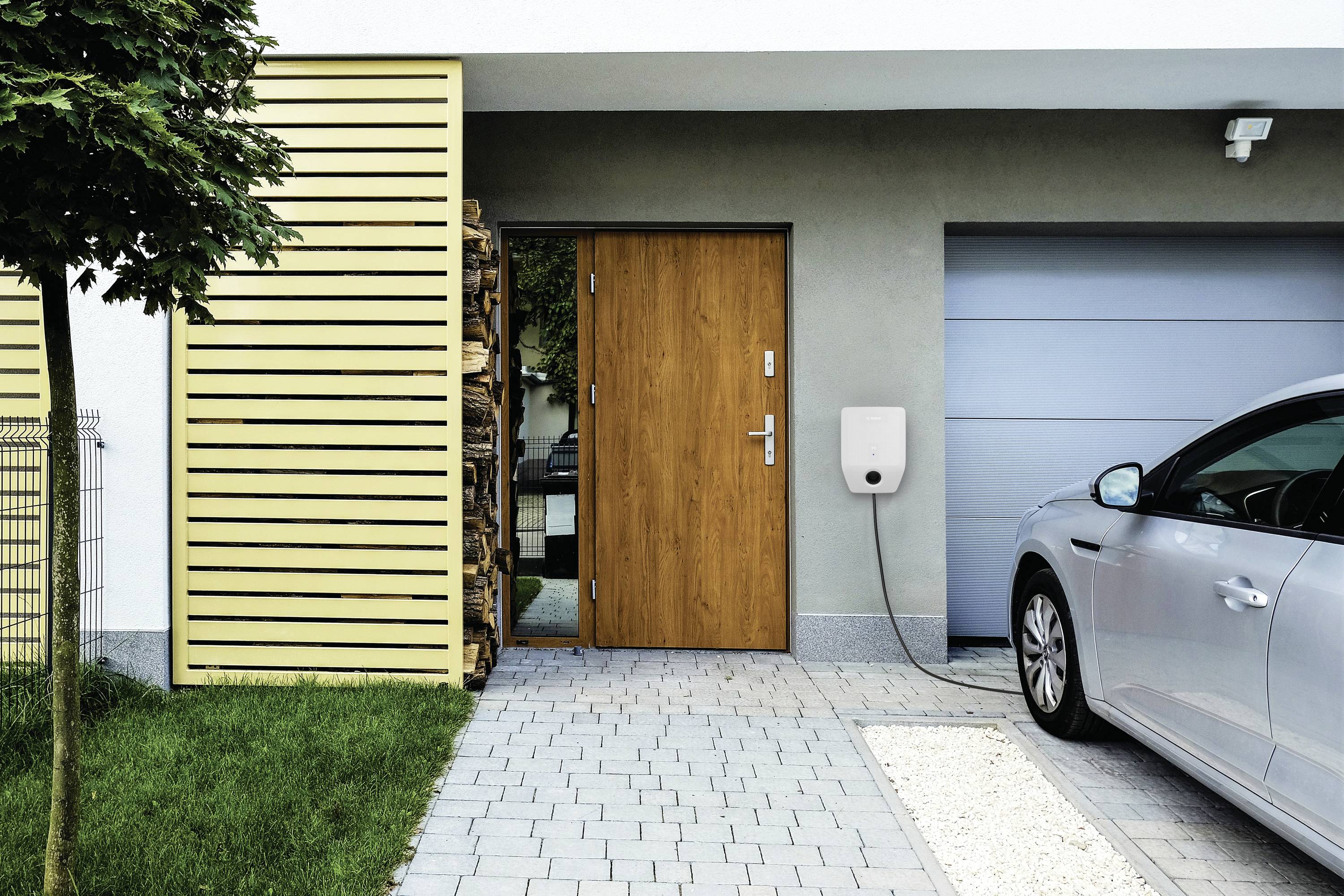 Ein graues Auto wird in einer Einfahrt neben einer Garage aufgeladen; das Ladekabel ist an eine Ladestation an der Wand angeschlossen.
