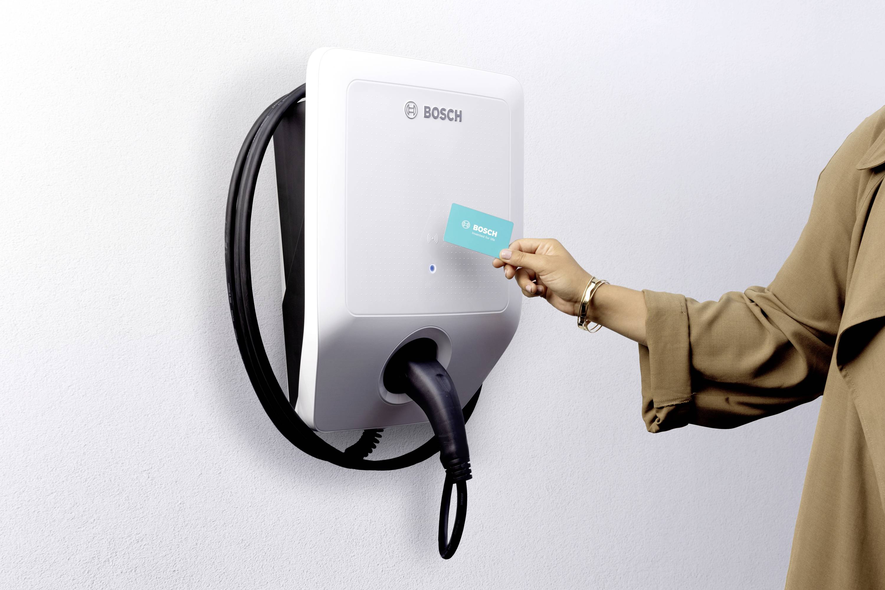 Eine Person hält eine Karte an ein Bosch-Ladegerät an der Wand. Das Gerät hat ein Kabel, das nach unten hängt.