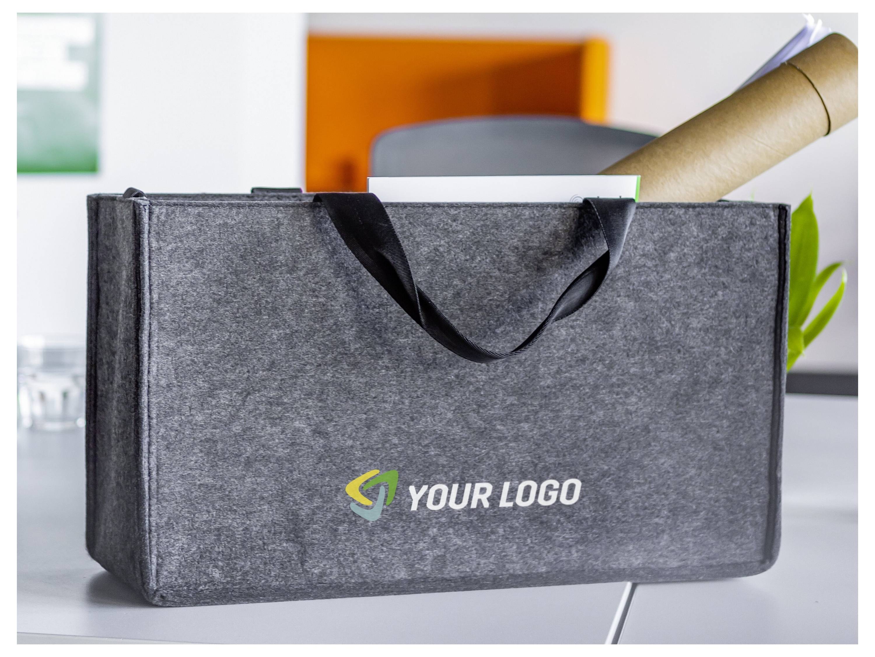 Graue isolierte Tragetasche mit schwarzen Griffen und "IHR LOGO"-Branding, auf einem Schreibtisch mit einer aufgerollten Zeitung oder einem Paket.