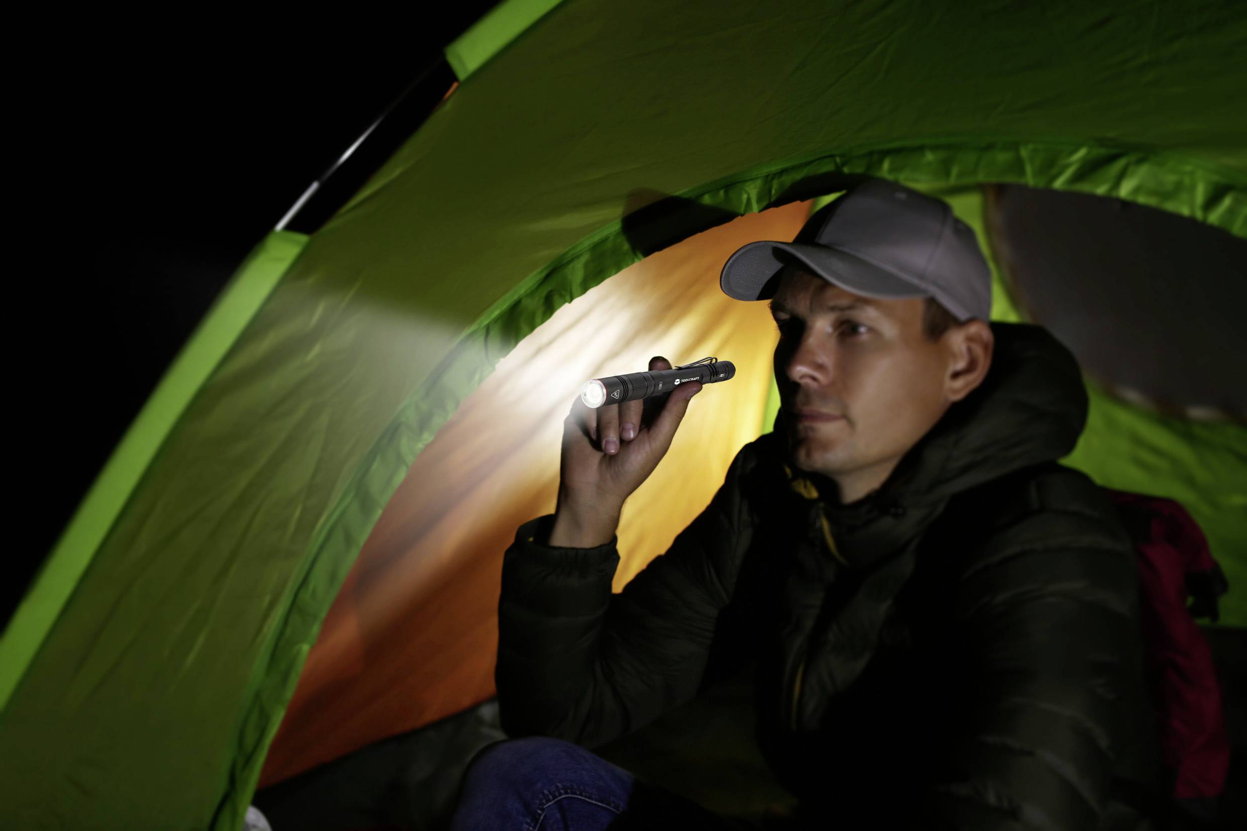 Person im Zelt sitzt bei Dunkelheit, hält Taschenlampe und schaut aufmerksam hinaus.