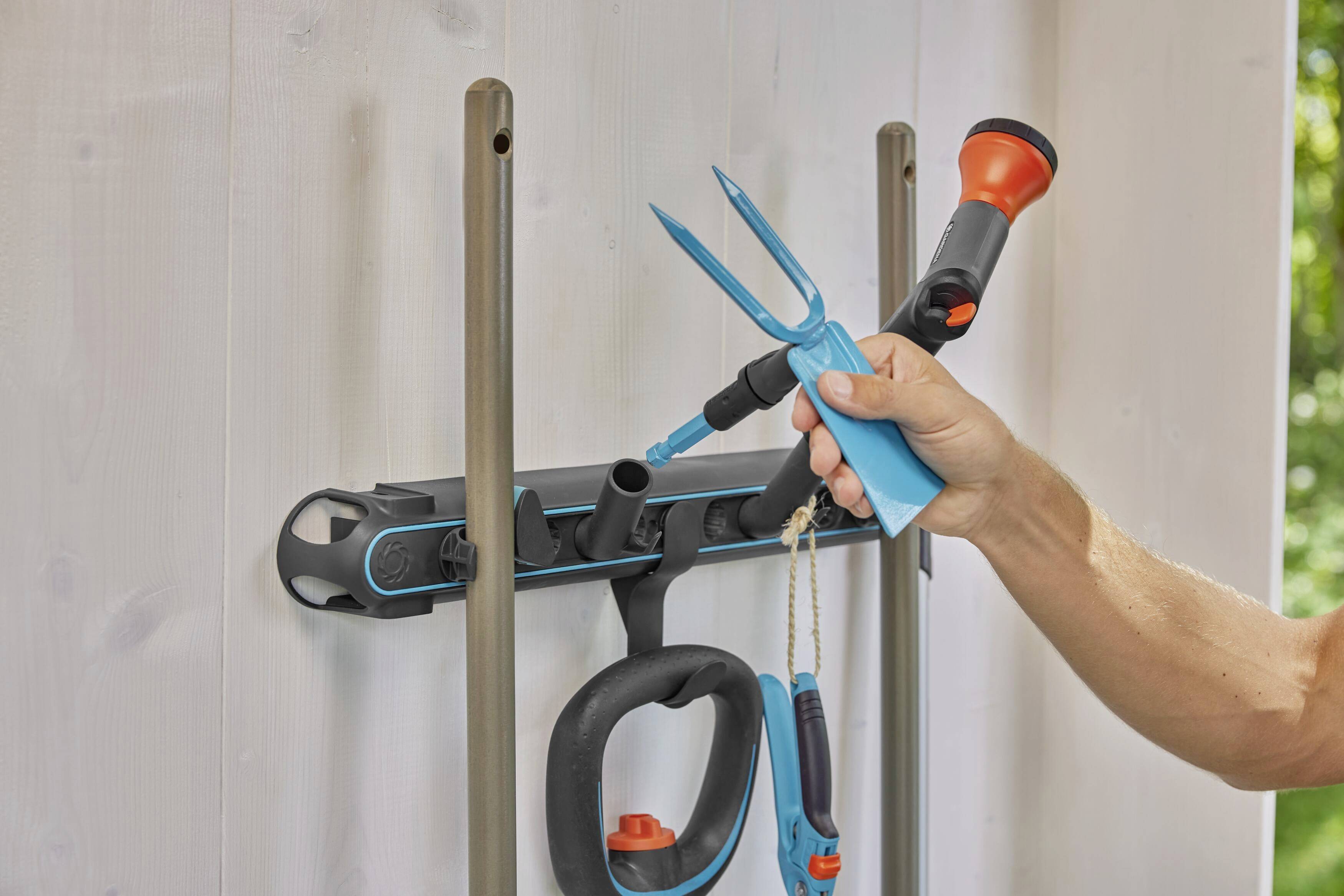 Eine Hand platziert eine blaue Gartenschere auf eine Werkzeughalterung an der Wand. Daneben hängen ein Schlauch und andere Gartengeräte.