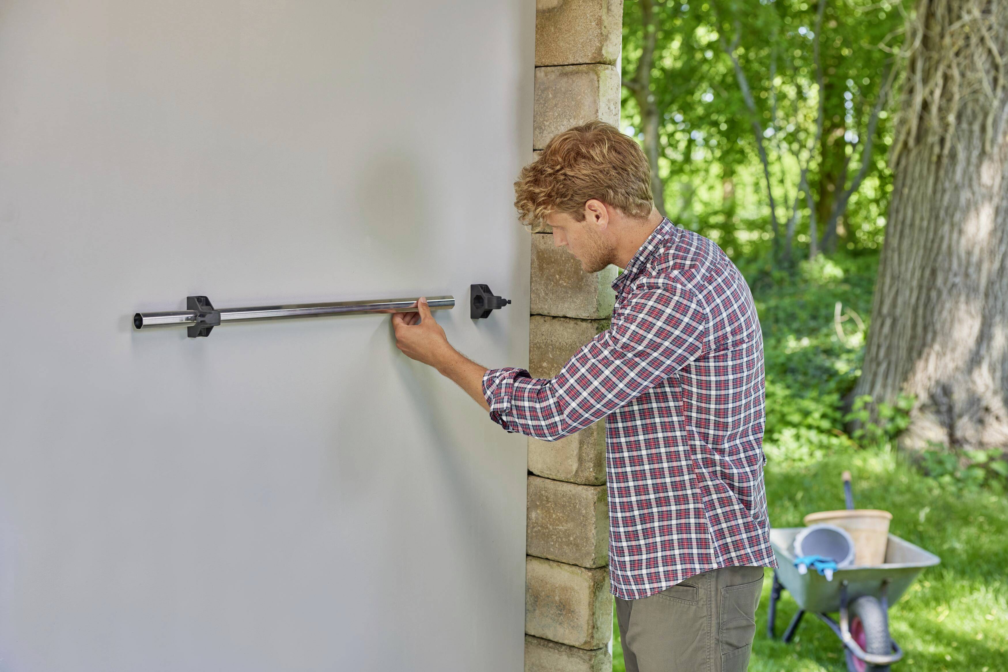 Ein Mann montiert eine Stange an eine rechteckige Wand im Freien. Neben ihm steht eine Schubkarre. Im Hintergrund sind Bäume zu sehen.