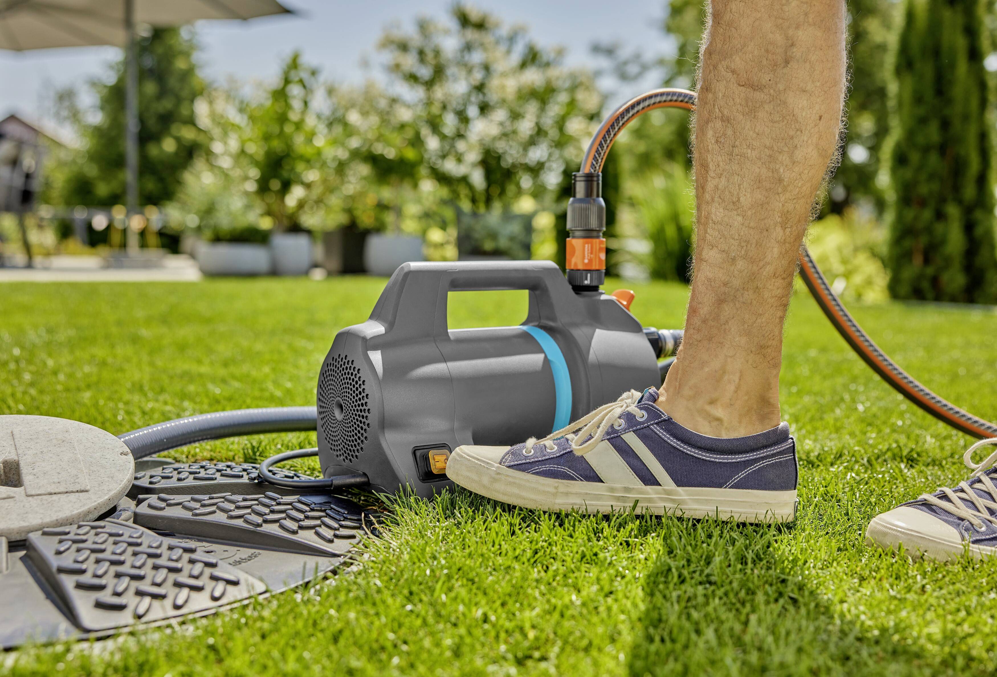 Eine Person tritt auf das Ventil einer schwarzen Gartenpumpe auf einem grünen Rasen, um Wasser zu pumpen. Ein Schlauch ist angeschlossen.