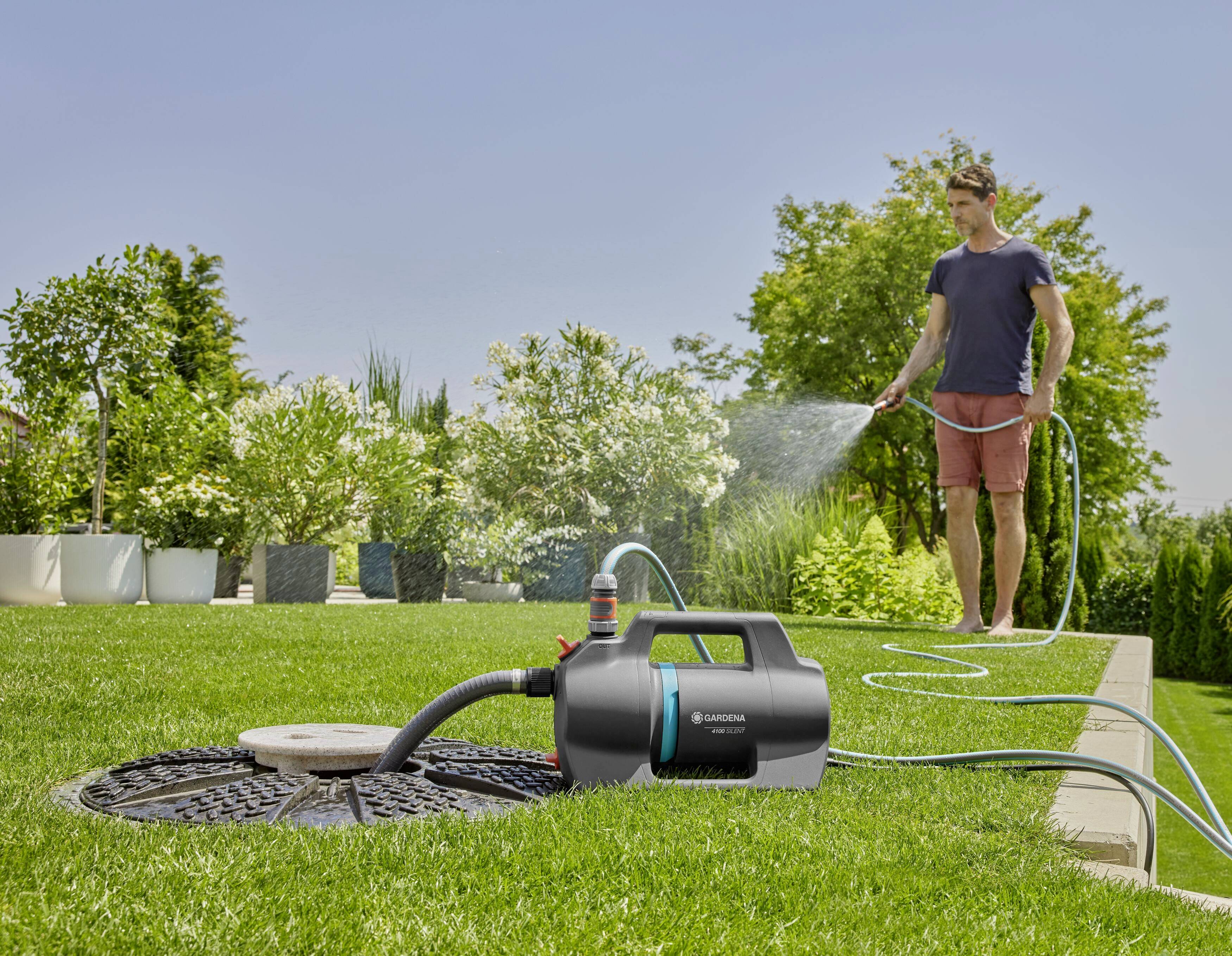 Ein Mann bewässert mit einem Gartenschlauch einen Rasen. Im Vordergrund steht eine elektrische Pumpe, die Wasser aus einem Brunnen zieht.