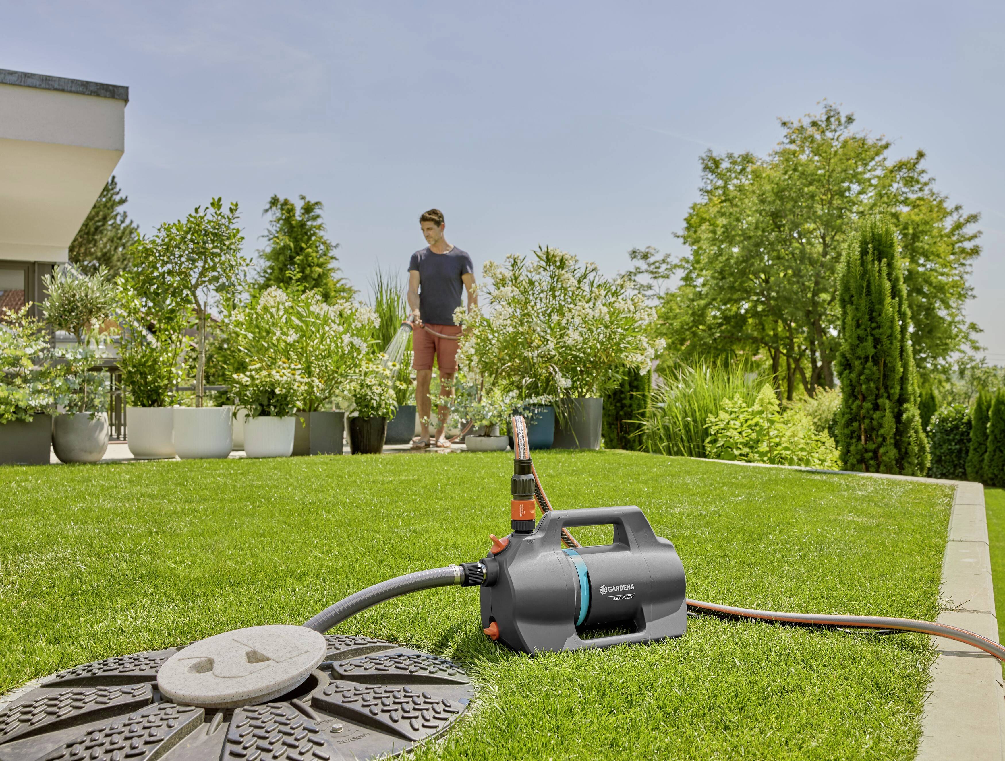 Eine Person bewässert einen grünen Rasen mit einem automatischen Bewässerungssystem im Garten. Im Hintergrund sind Pflanzen und Bäume zu sehen.