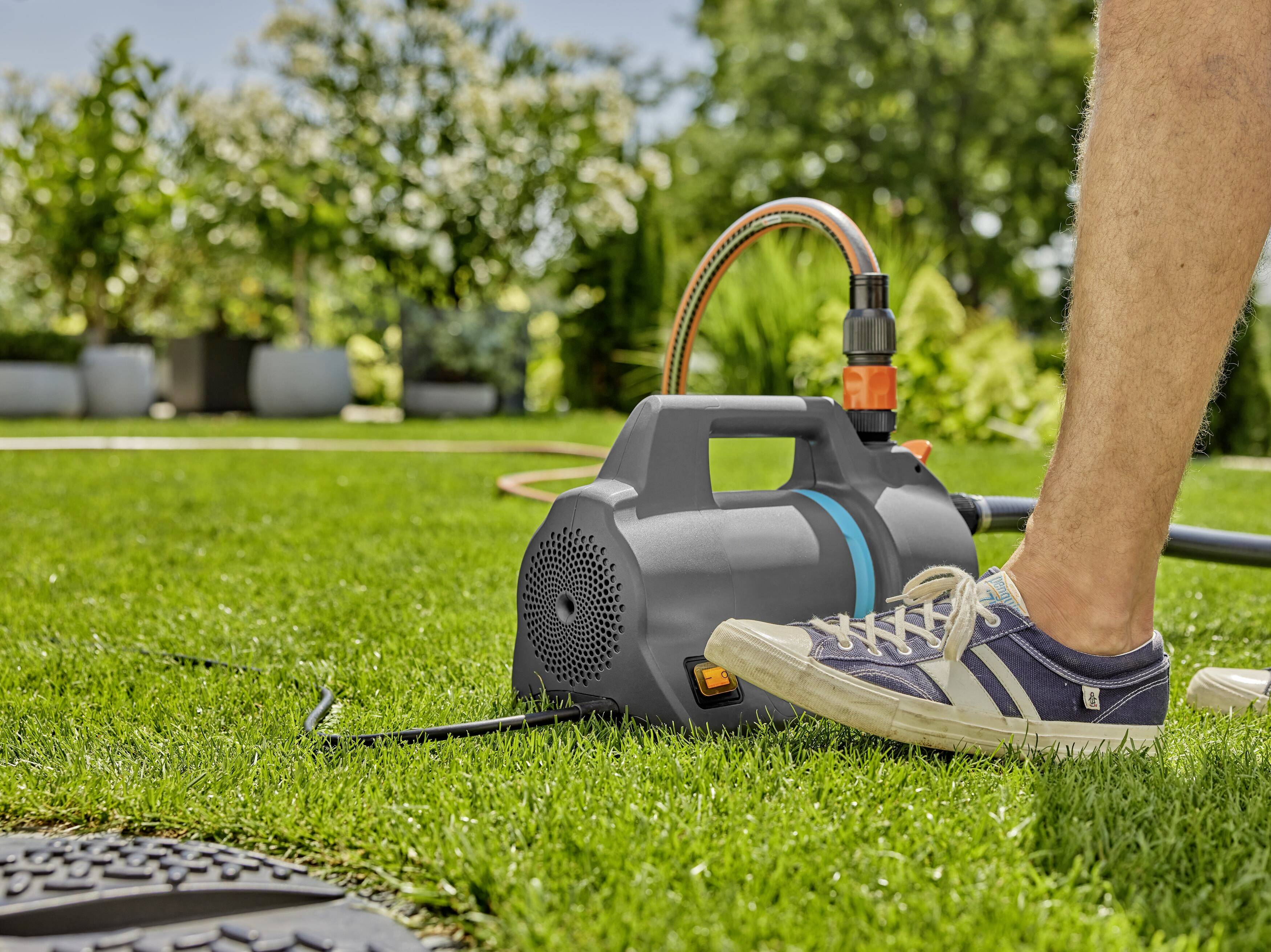 Eine Person schaltet mit dem Fuß eine elektrische Wasserpumpe im Garten ein. Die Pumpe hat einen Wasserschlauch angeschlossen.
