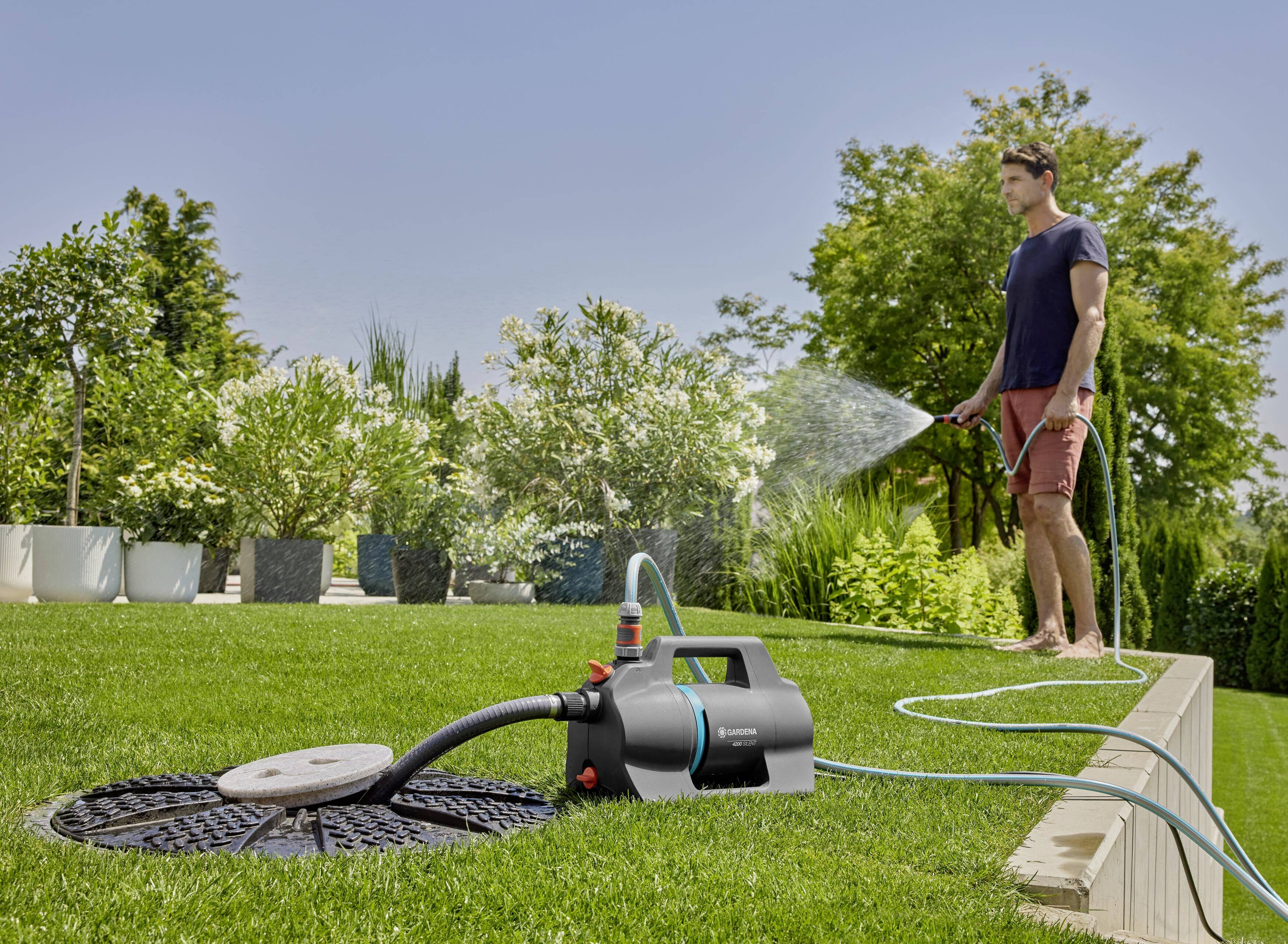 Ein Mann bewässert einen Rasen mit einem Schlauch, der an eine Gartenpumpe angeschlossen ist. Im Hintergrund sind Bäume und Büsche sichtbar.
