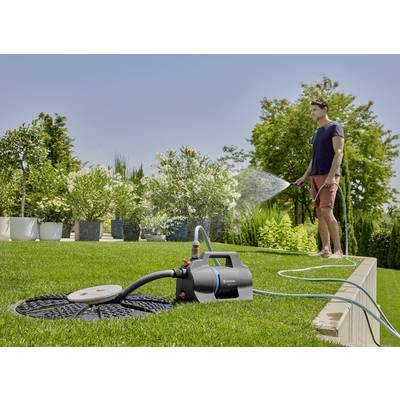 Ein Mann bewässert einen Rasen mit einem Schlauch, der an eine Gartenpumpe angeschlossen ist. Im Hintergrund sind Bäume und Büsche sichtbar.