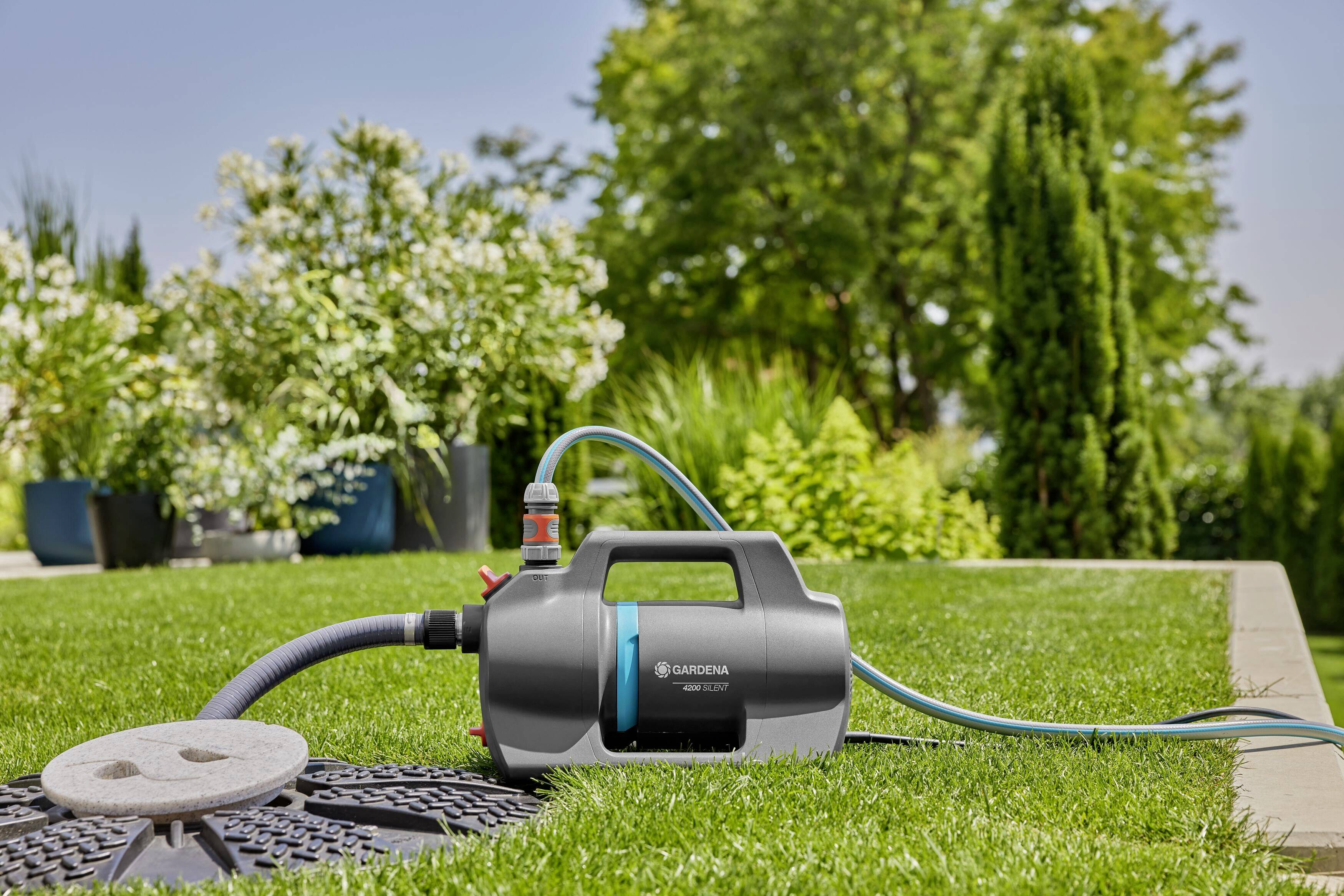 Eine Gartenpumpe auf einem Rasen, umgeben von Pflanzen, ist mit einem Schlauch verbunden und bereit zur Wasserbewässerung.