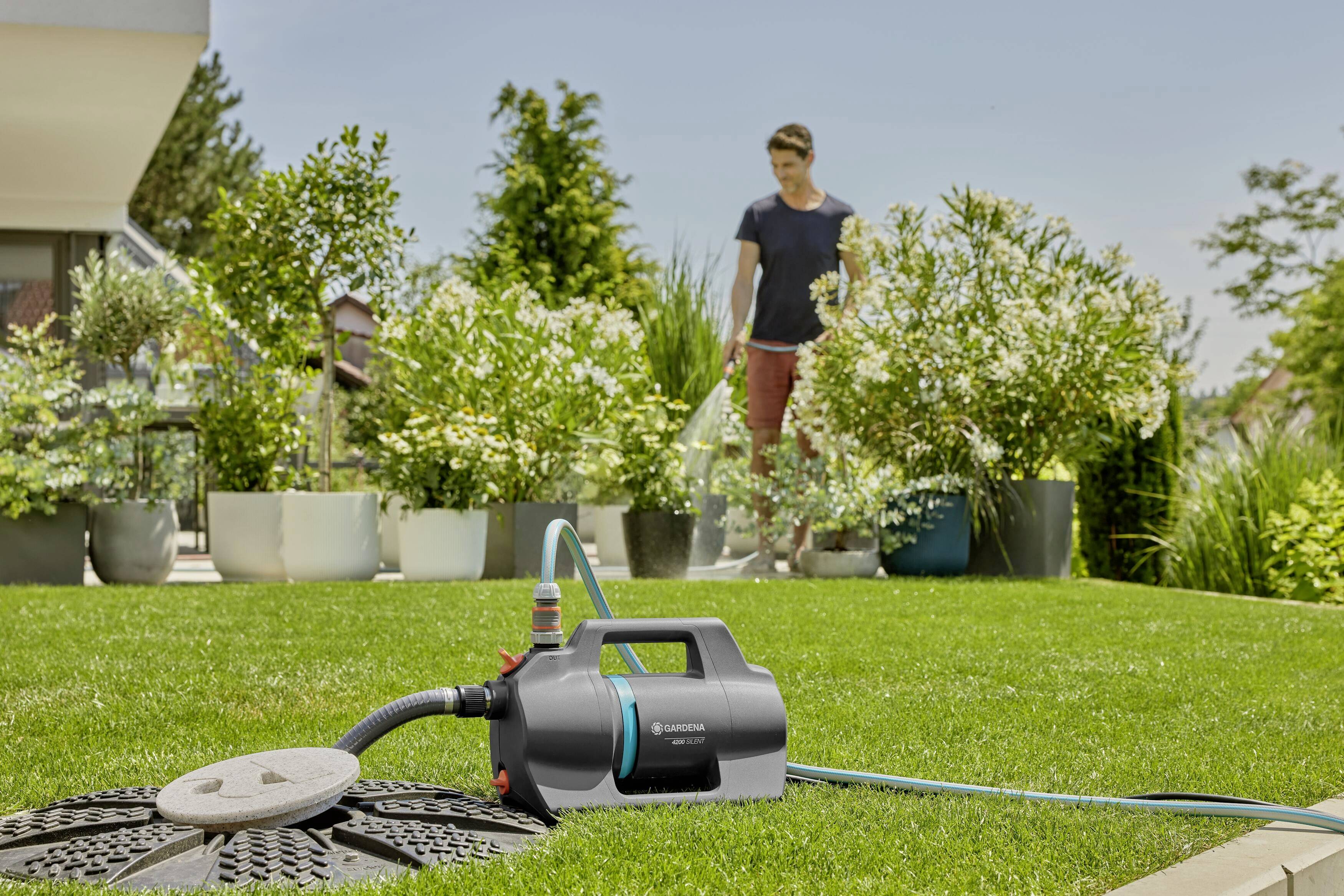 Eine Person im Garten, im Hintergrund; Vordergrund: Gartenpumpe mit angeschlossenem Schlauch, umgeben von Pflanzen und Rasen.
