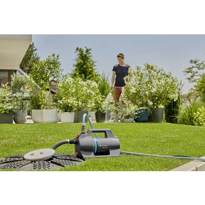Eine Person im Garten, im Hintergrund; Vordergrund: Gartenpumpe mit angeschlossenem Schlauch, umgeben von Pflanzen und Rasen.