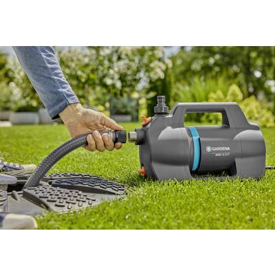 Eine Hand schließt einen Schlauch an eine Gardena Gartenpumpe auf einem Rasen an. Im Hintergrund sind Bäume und Sträucher sichtbar.