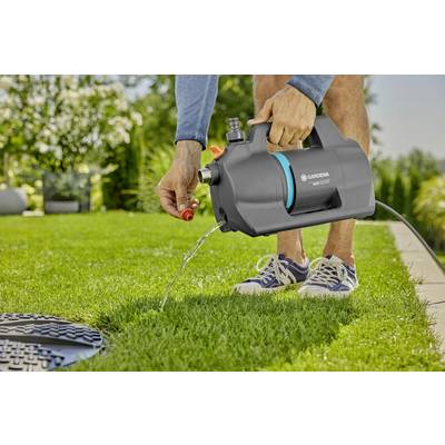 Eine Person hält eine schwarze Gartenpumpe über einem gepflegten Rasen. Wasser fließt aus der Pumpe, während im Hintergrund Pflanzen zu sehen sind.