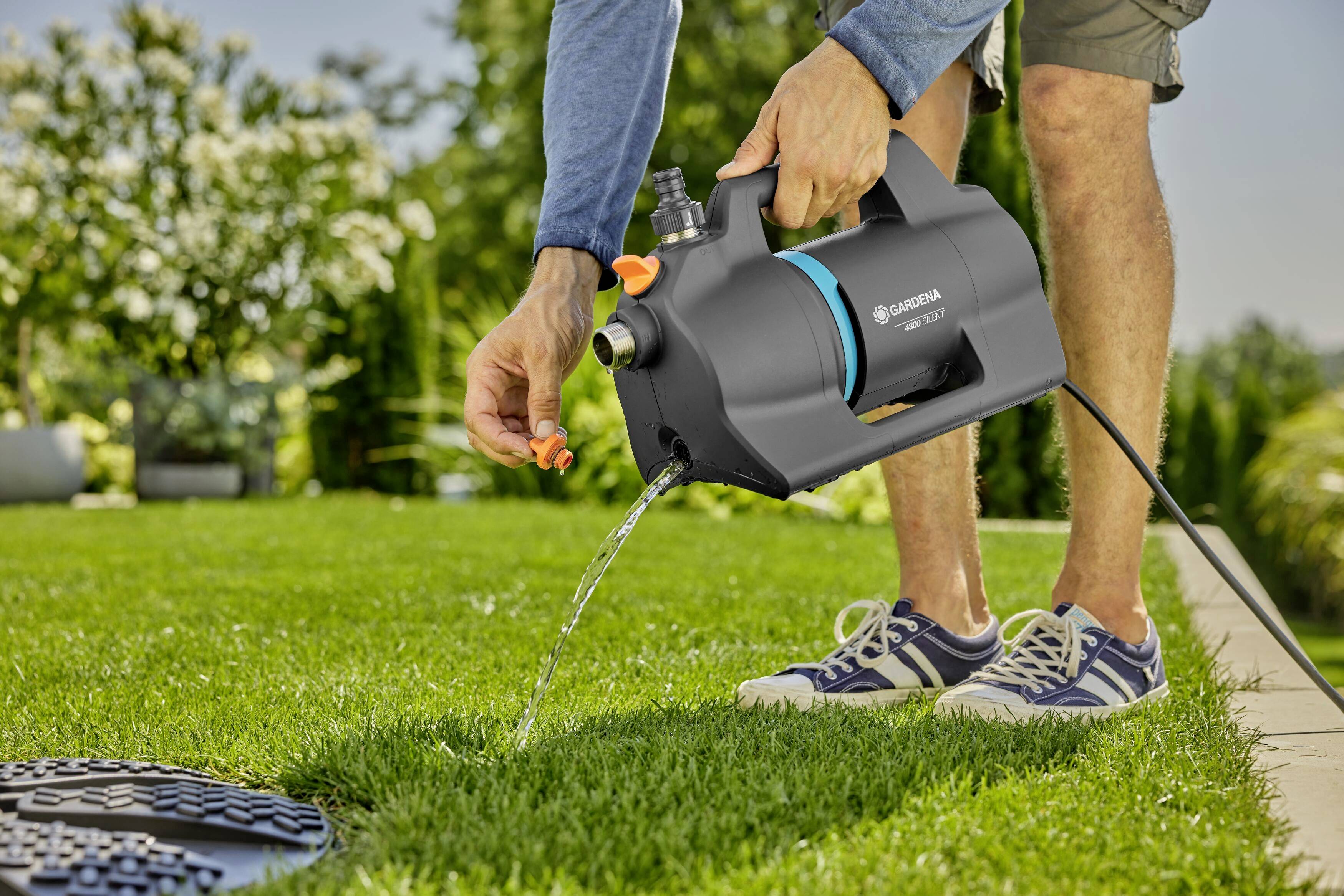 Eine Person wässert den Rasen mit einer tragbaren Pumpe im Garten. Im Hintergrund sind Pflanzen und Bäume zu sehen.