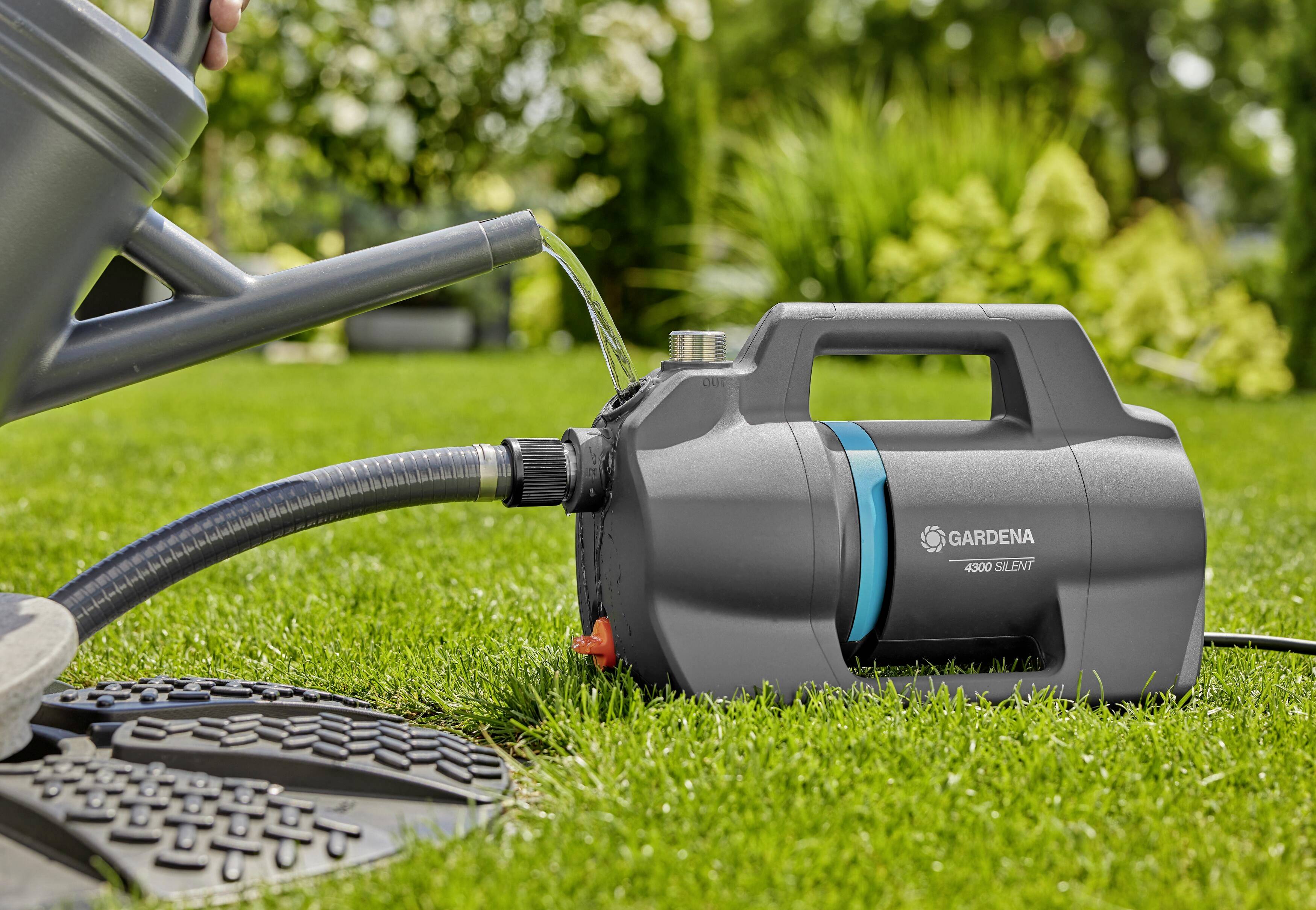 Eine Gartenpumpe bewässert einen Rasen. Ein Wasserschlauch transportiert das Wasser aus einem beleuchteten Gullydeckel.