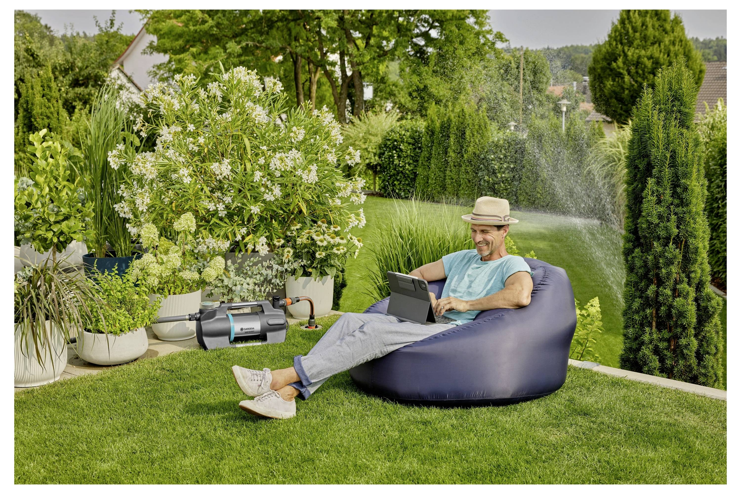 Eine Person mit einem Hut sitzt auf einem Sitzsack im Garten und arbeitet an einem Laptop. Ganz in der Nähe bewässert ein Sprinkler den grünen Rasen und die Pflanzen.