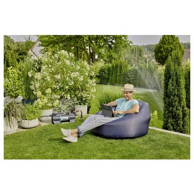Eine Person mit einem Hut sitzt auf einem Sitzsack im Garten und arbeitet an einem Laptop. Ganz in der Nähe bewässert ein Sprinkler den grünen Rasen und die Pflanzen.