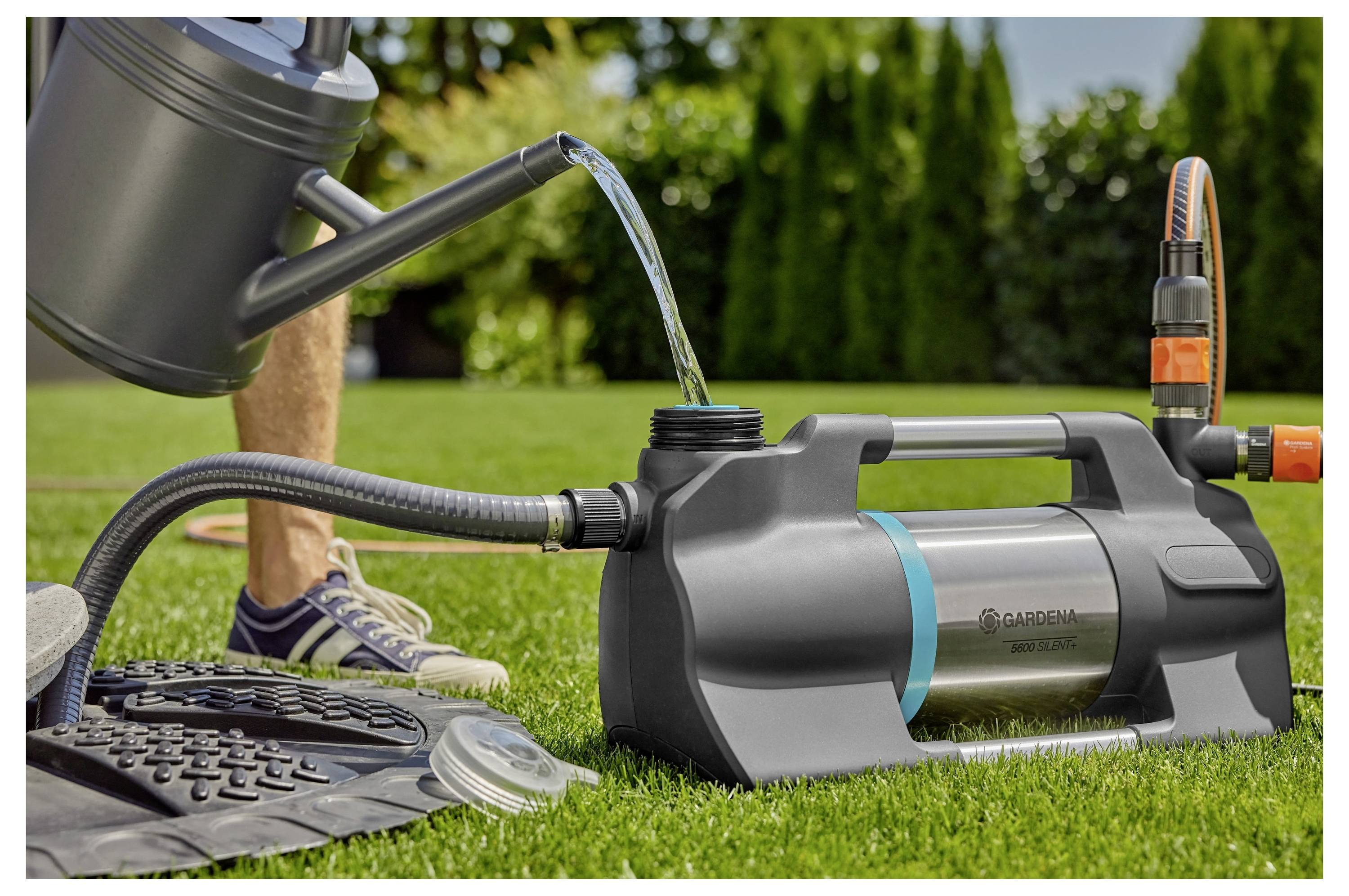 „Eine Gartenpumpe, die Wasser aus einem Reservoir in eine Gießkanne auf einem Rasen umleitet, mit einer Person im Hintergrund, die Turnschuhe trägt."