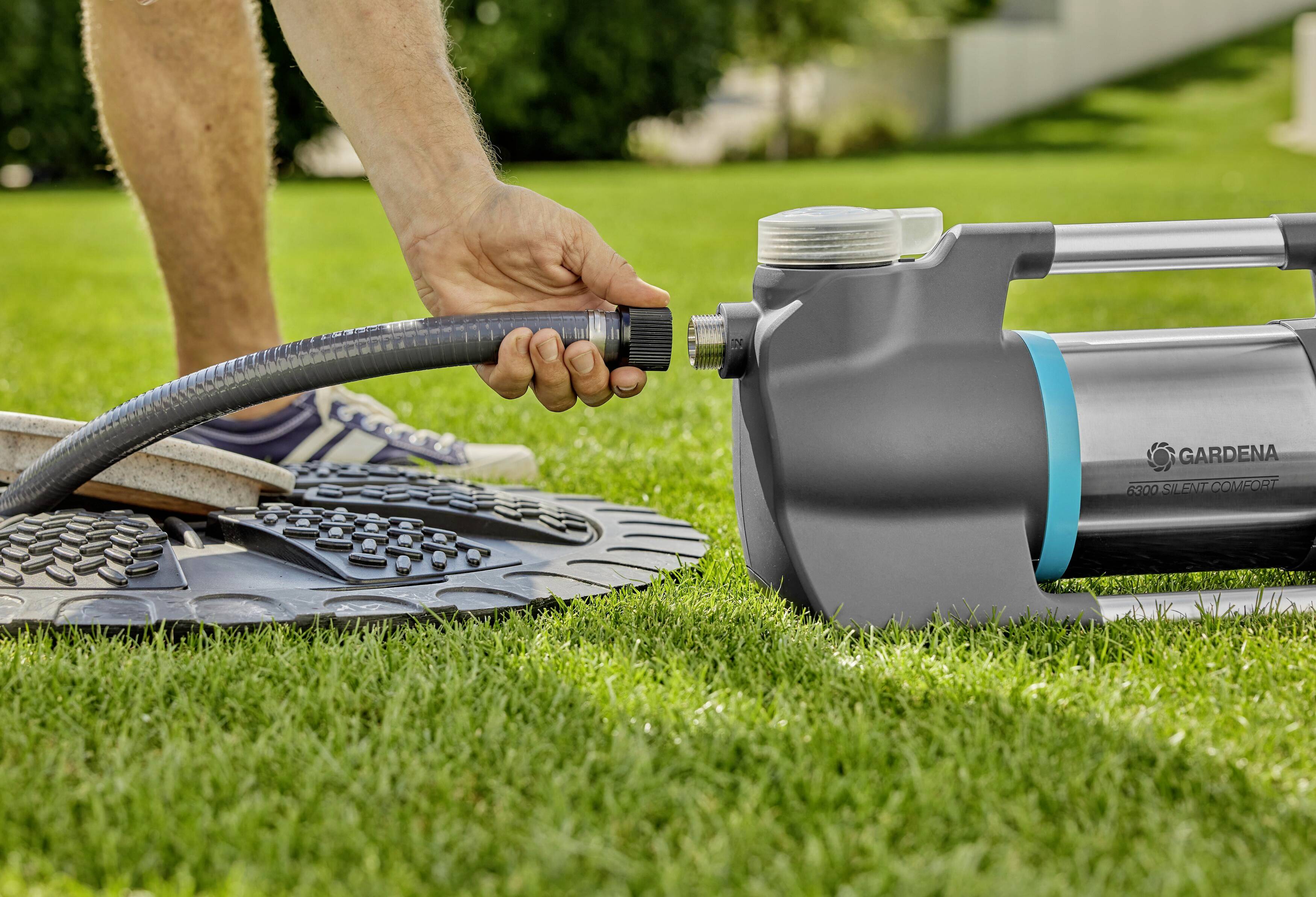 Eine Person verbindet einen Gartenschlauch mit einer Wasserpumpe auf einem Rasen. Im Hintergrund sind Gartenpflanzen zu sehen.