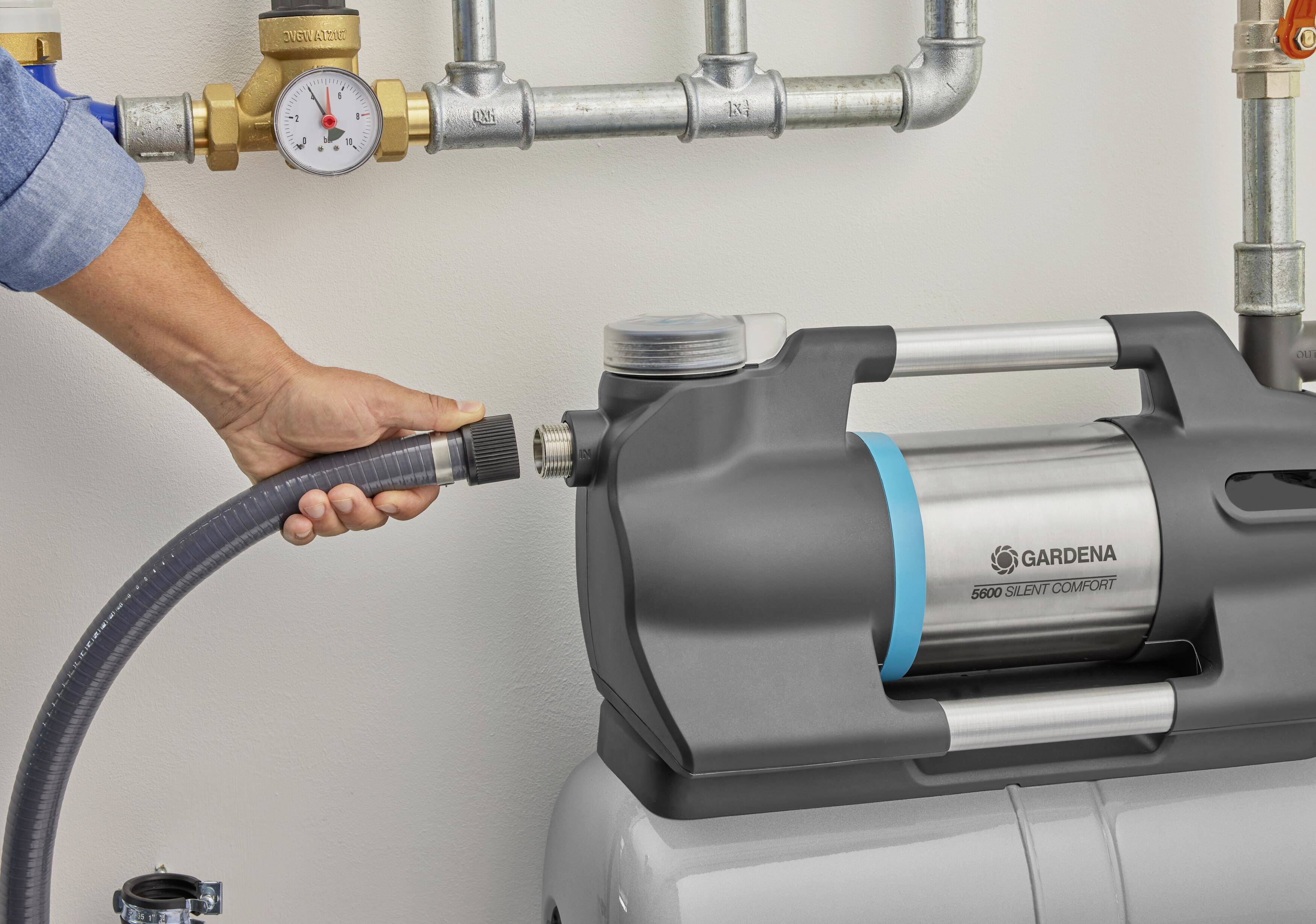 Eine Person verbindet einen Gartenschlauch mit einer Wasserpumpe in einer Werkstatt. Oben sind Rohrleitungen mit einem Manometer sichtbar.