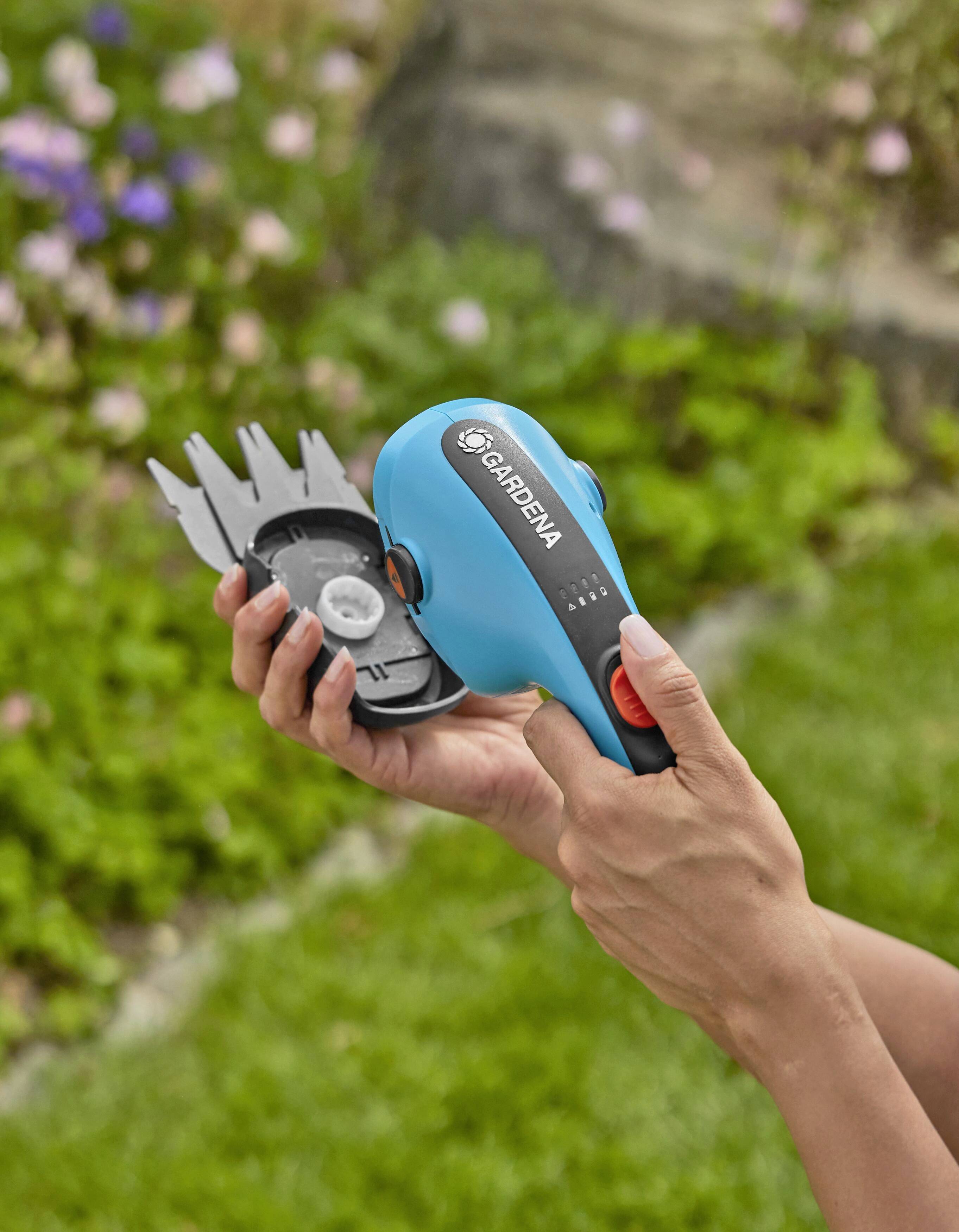 Person hält eine blaue elektrische Hecke- und Rasenkantenschere in einem Garten. Hintergrund zeigt unscharfe Blumen und Rasen.