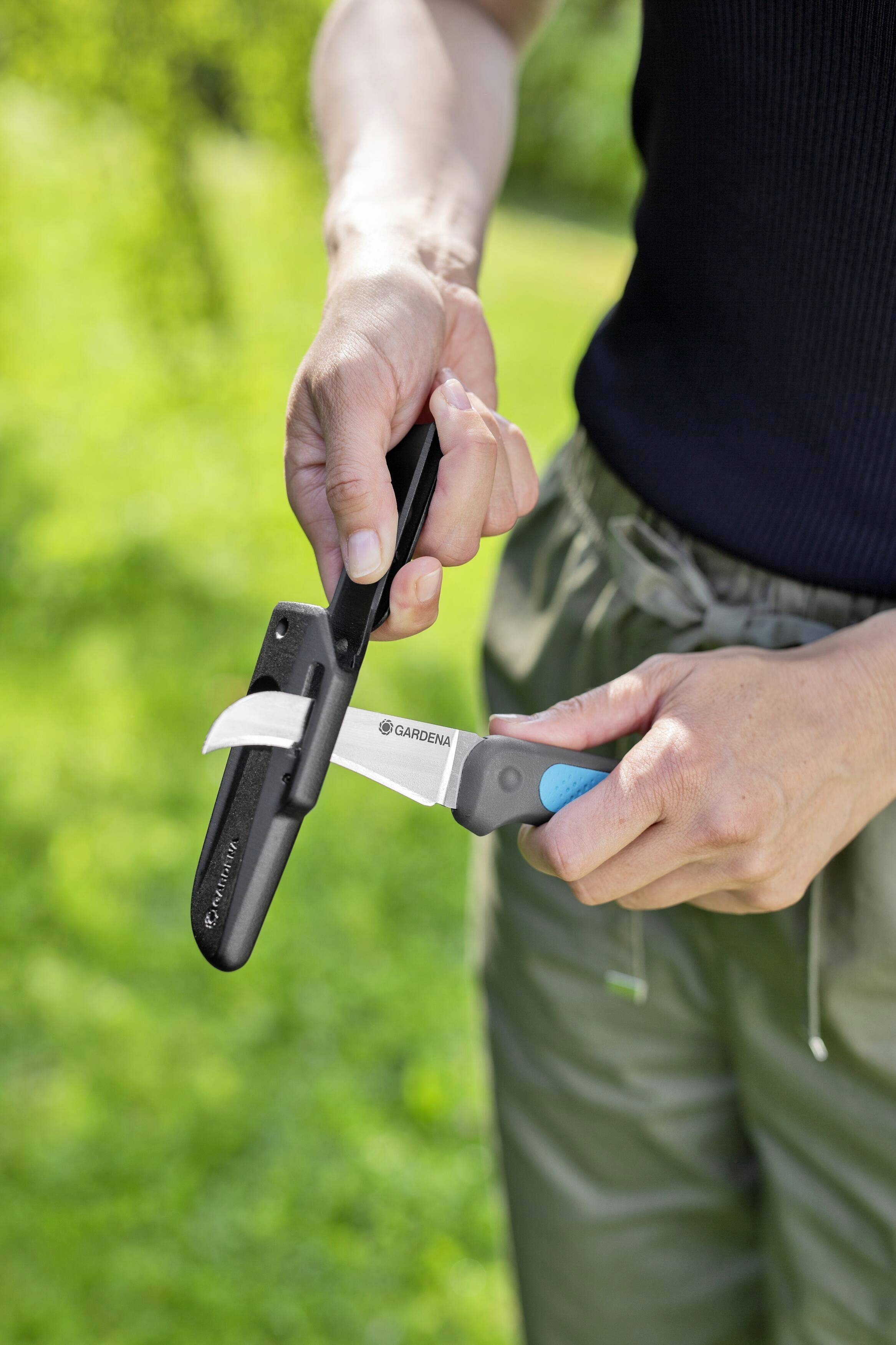 Person schleift ein Messer mit einem Hand-Schärfer im Freien. Sonniges Wetter, grüner Hintergrund.