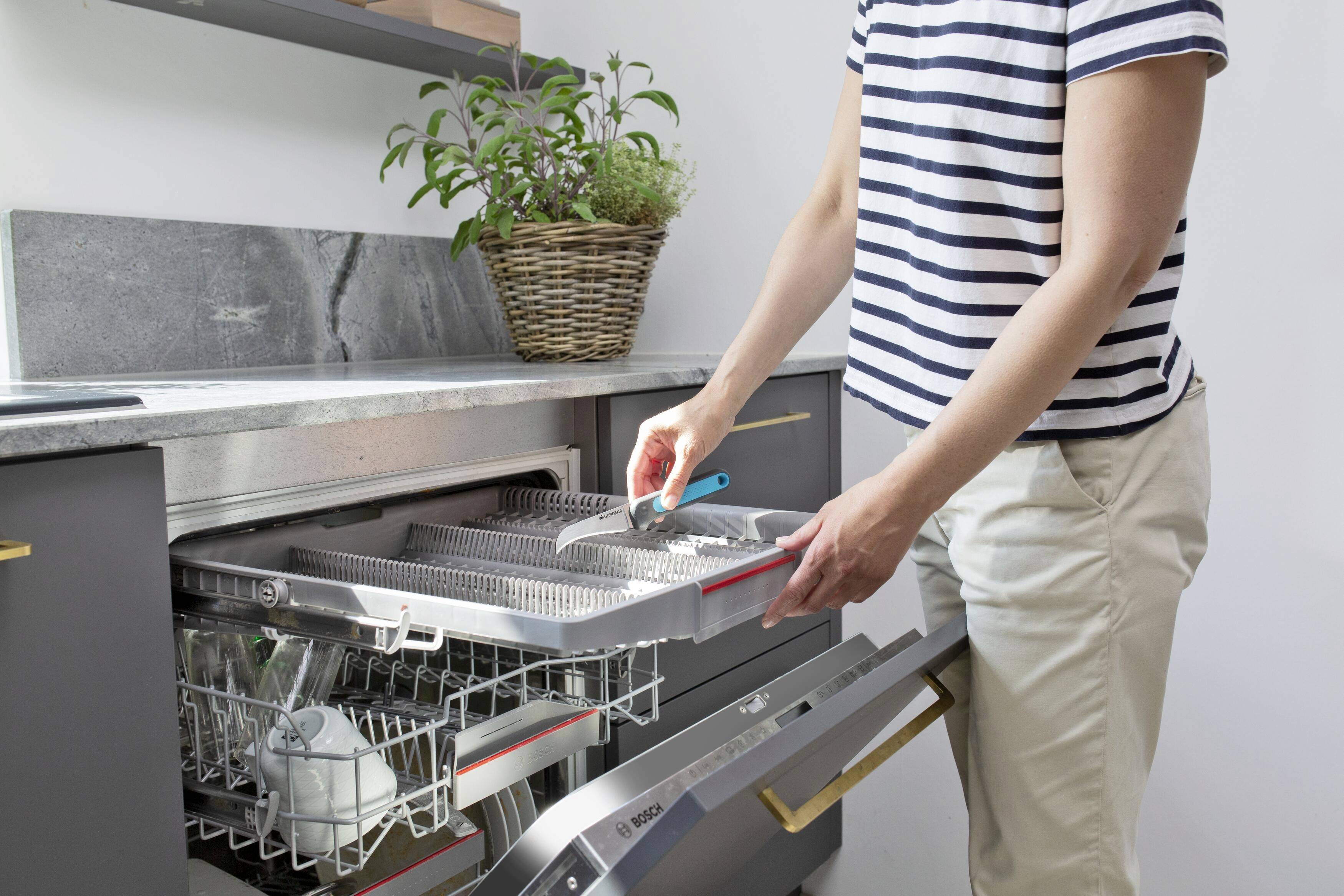 Eine Person räumt eine Spülmaschine ein, hält Besteck in der Hand. Im Hintergrund steht ein Korb mit Kräutern auf der Arbeitsplatte.