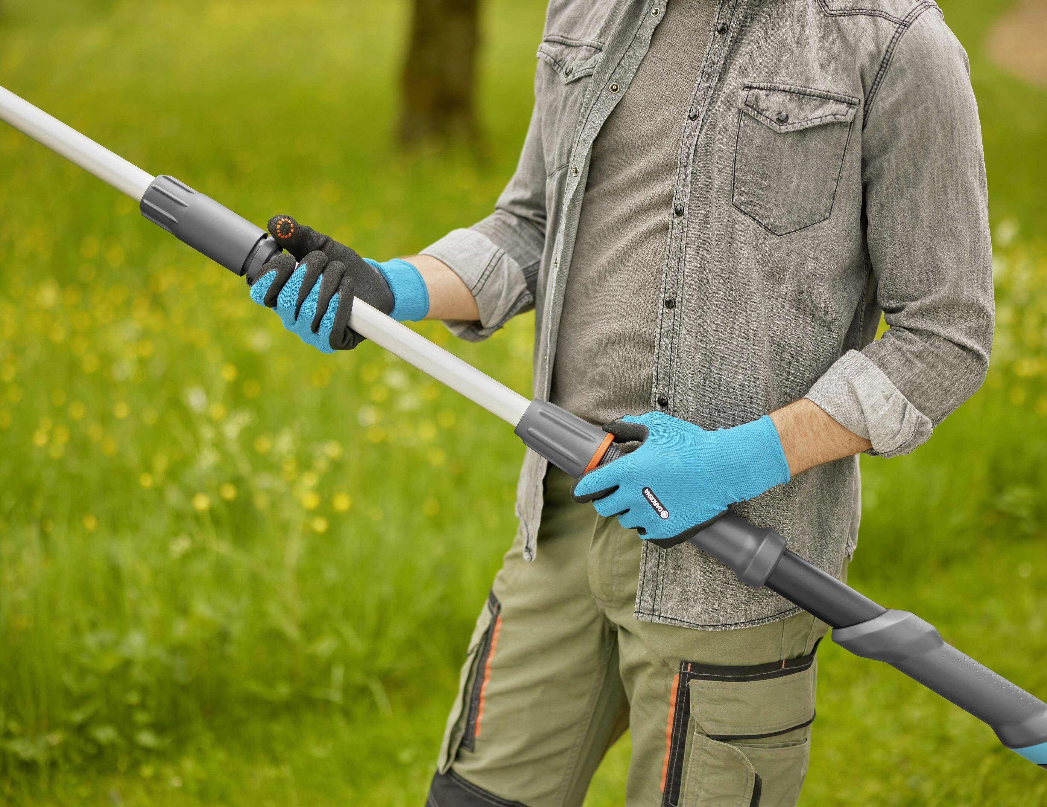 Person hält einen Teleskop-Gartenschlauch in einem Garten. Person trägt Handschuhe. Hintergrund zeigt grünes Gras und Bäume.