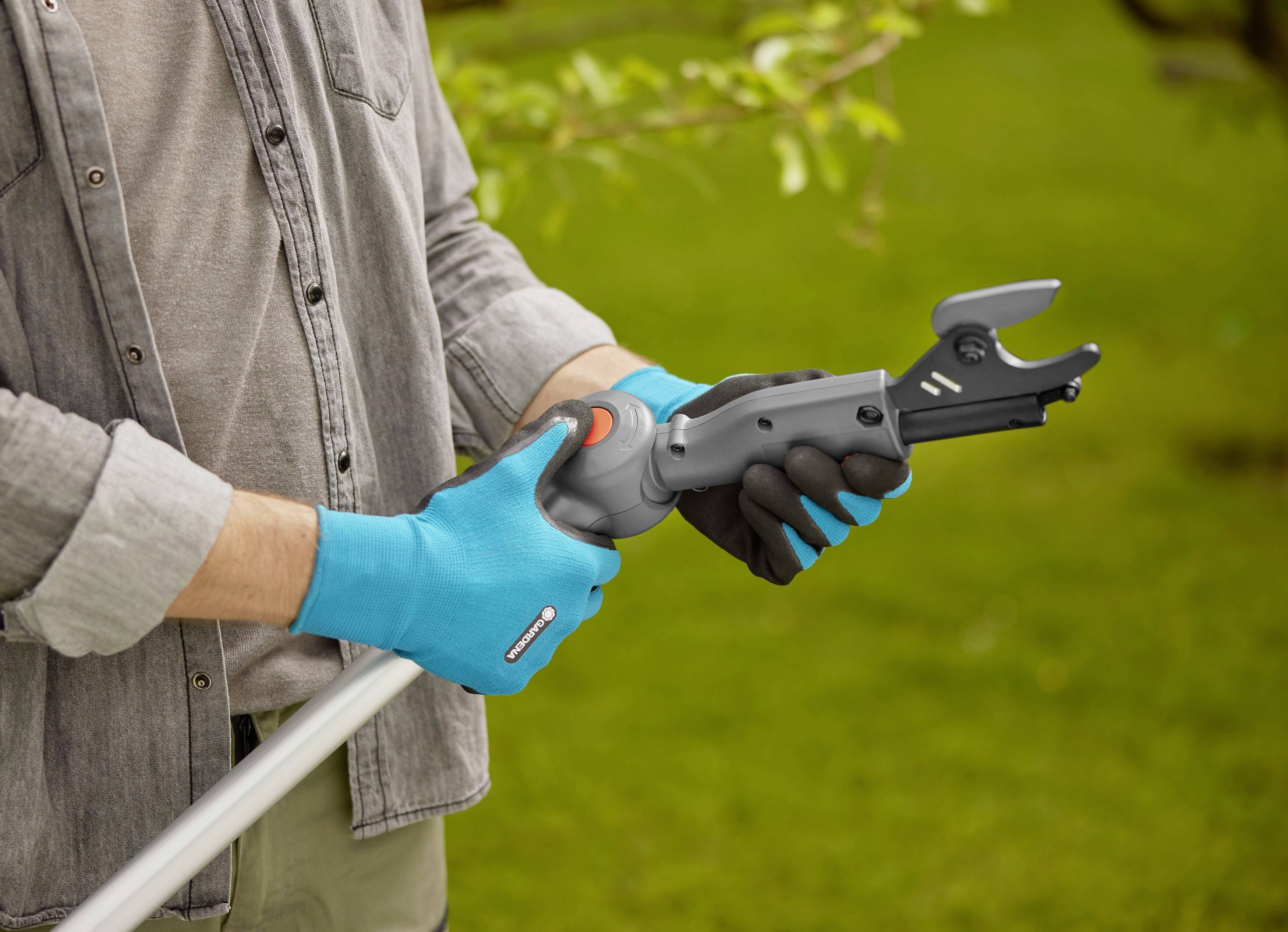 Person schneidet mit einer Astschere im Garten. Handschuhe schützen die Hände. Grünflächen und Bäume sind im Hintergrund sichtbar.