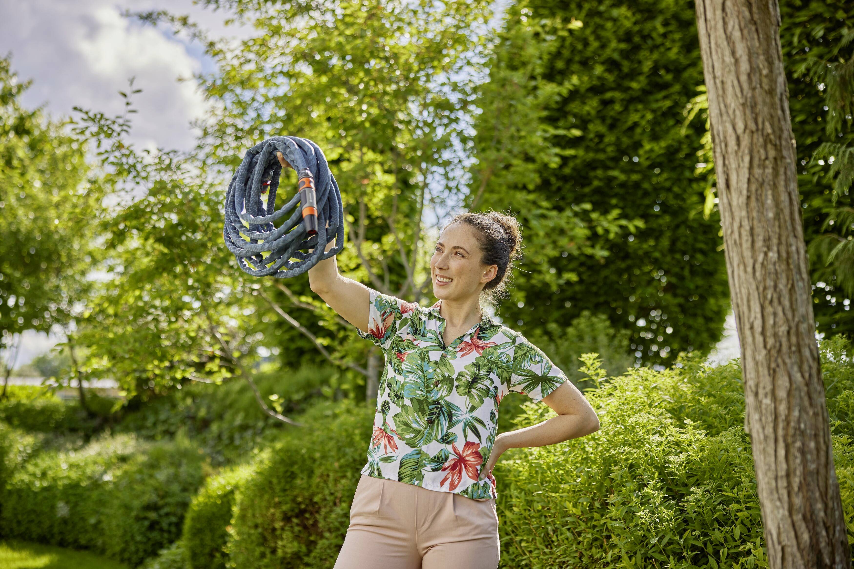 Person hält einen zusammengewickelten Gartenschlauch in einem sonnigen Garten. Umgeben von grünen Büschen und Bäumen.