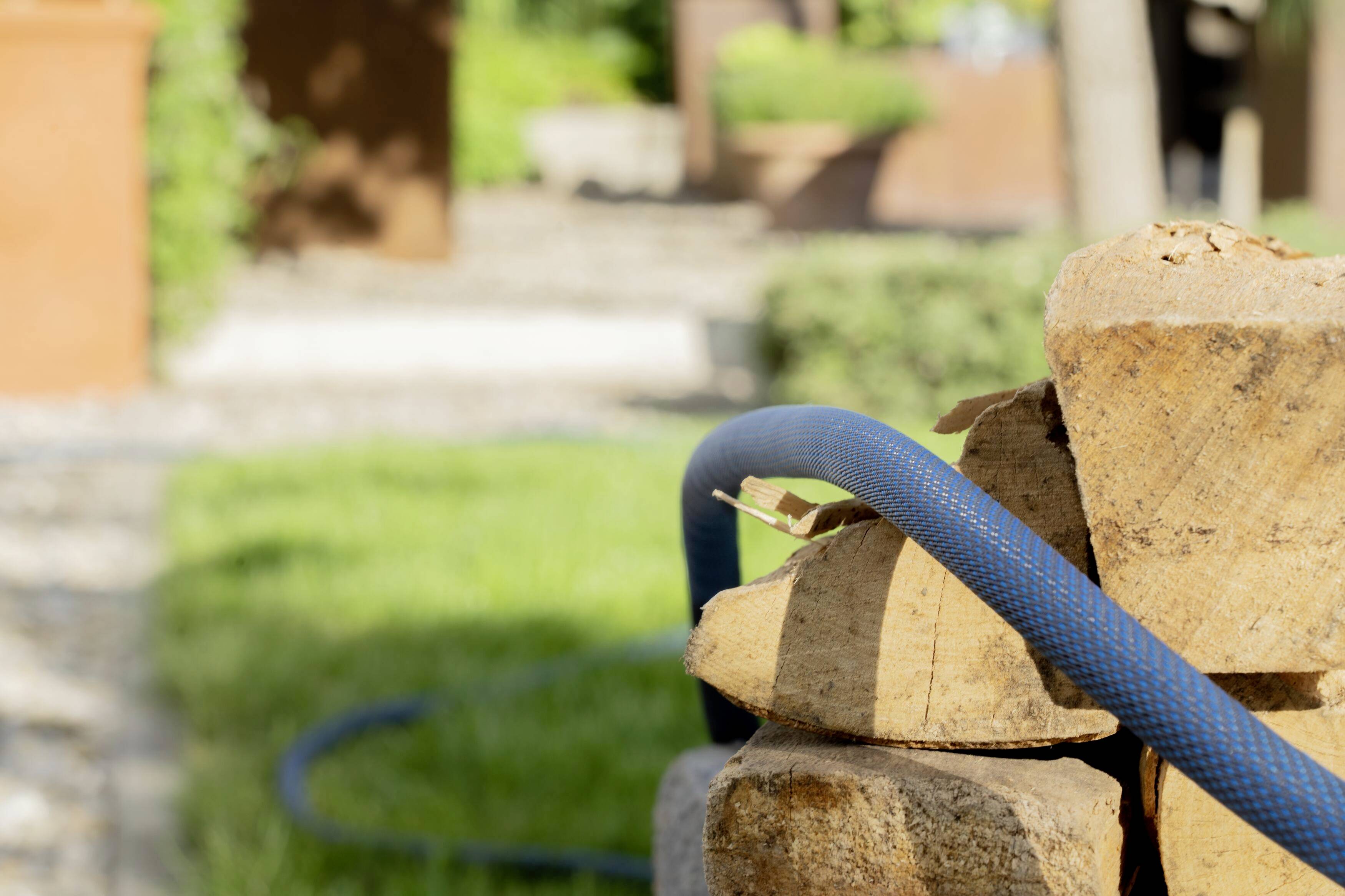 Ein blauer Gartenschlauch ruht auf einem Stapel Holzscheite im sonnigen Garten, umgeben von Gras und verschwommenen Hintergrundstrukturen.