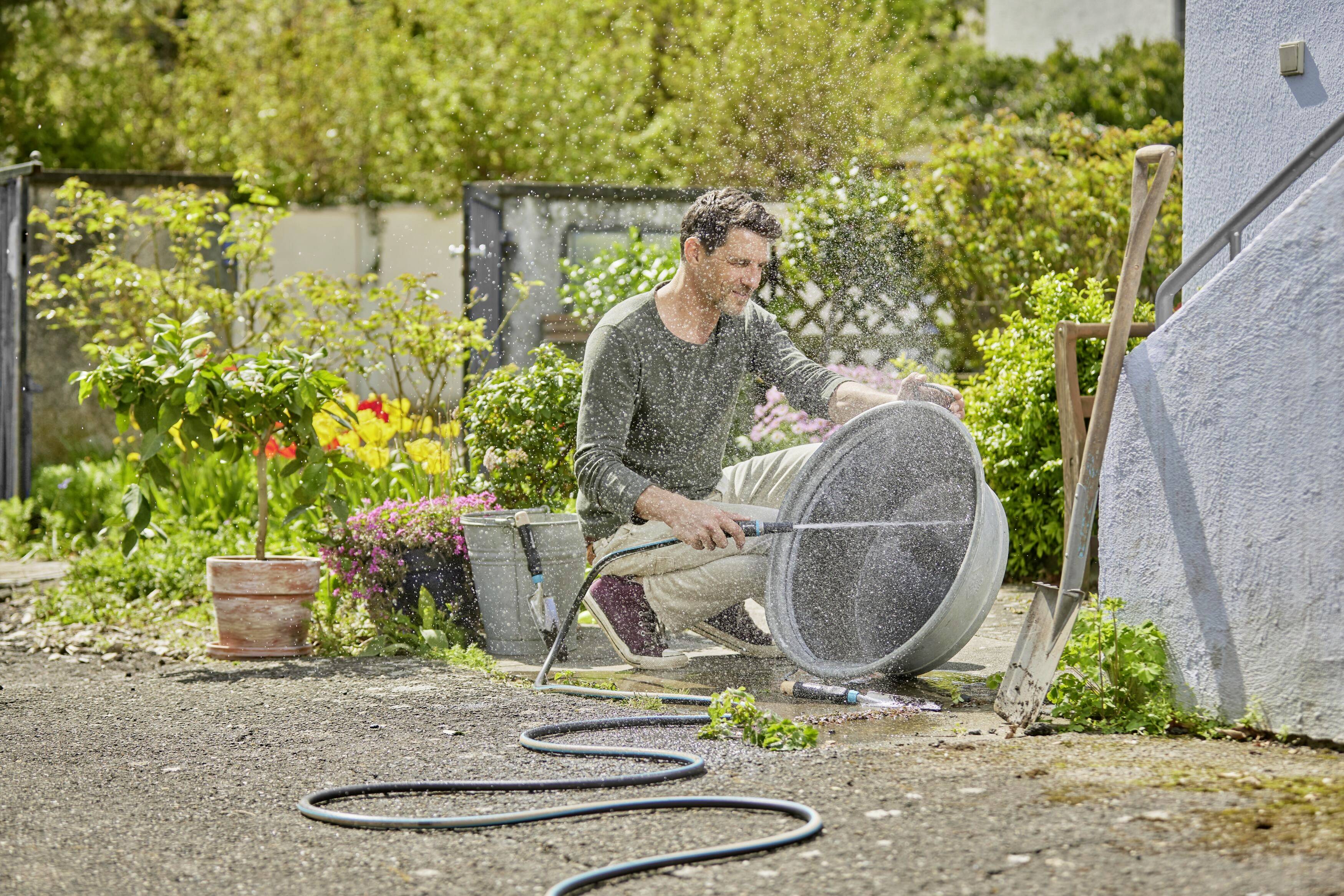 Ein Mann reinigt im Garten ein großes rundes Sieb mit einem Wasserschlauch. Im Hintergrund sind Pflanzen und eine Gartenmauer zu sehen.