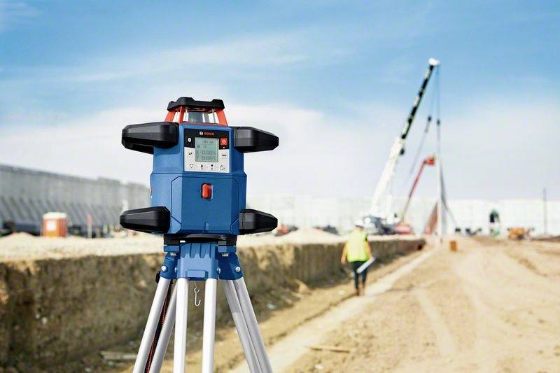 Ein Nivelliergerät steht im Vordergrund einer Baustelle. Im Hintergrund arbeiten Arbeiter und Kräne an einem Gebäude.