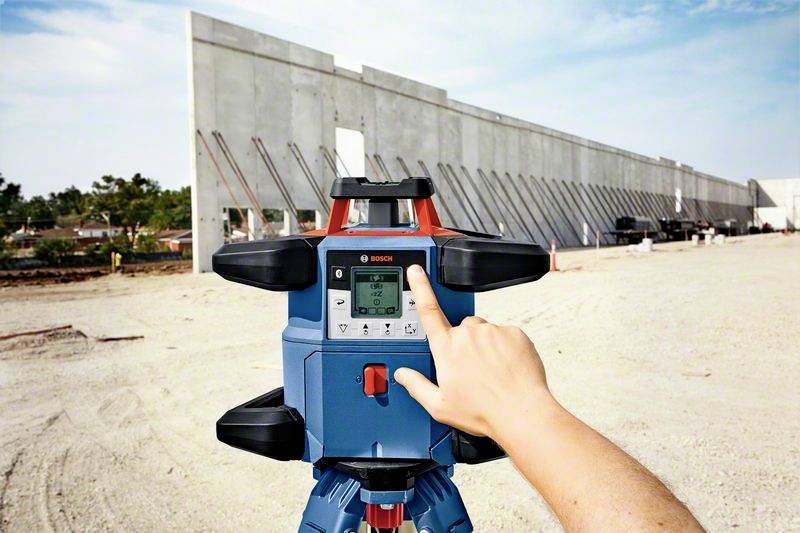 Eine Hand bedient ein Laser-Nivelliergerät auf einer Baustelle mit im Hintergrund stehenden Betonwänden.