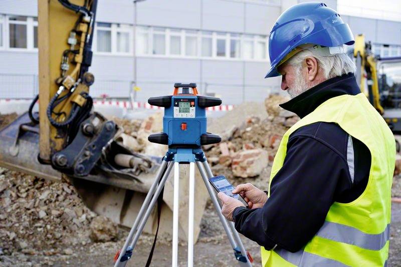 Ein Arbeiter mit Schutzhelm und Weste bedient ein Vermessungsgerät auf einer Baustelle. Im Hintergrund ist ein Bagger zu sehen.