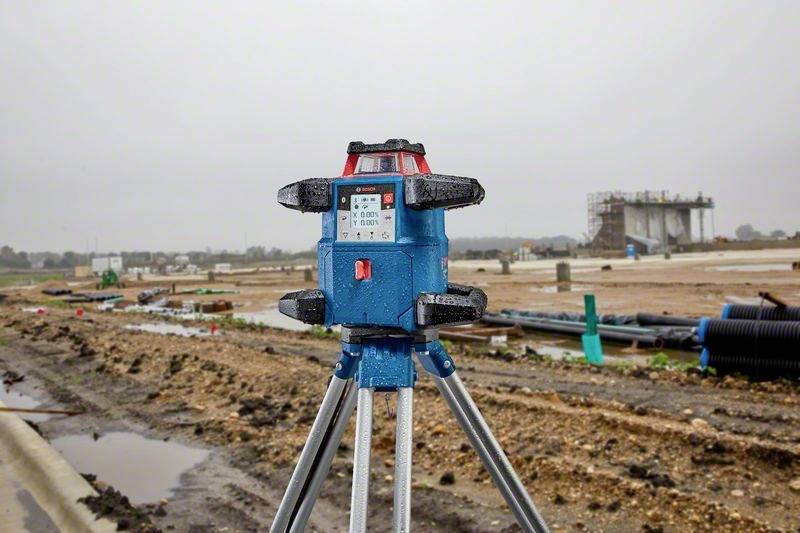 Ein Laser-Nivelliergerät steht auf einer Baustelle, umgeben von Rohren und Baumaterialien, bei bewölktem Himmel. Im Hintergrund ist ein Gebäude im Bau.