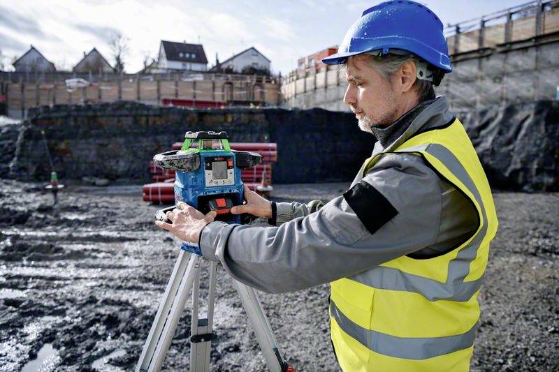 Arbeiter in Baukleidung benutzt ein Vermessungsgerät auf einer Baustelle, um Bodenunebenheiten zu messen. Häuser im Hintergrund.