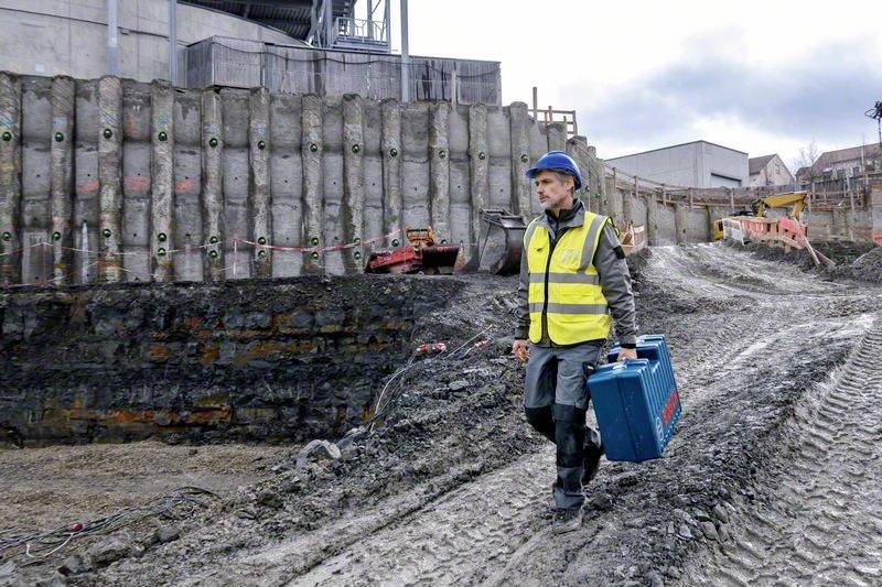 Ein Bauarbeiter in Sicherheitsausrüstung und Helm trägt einen blauen Werkzeugkoffer über eine schlammige Baustelle mit Betonwänden.