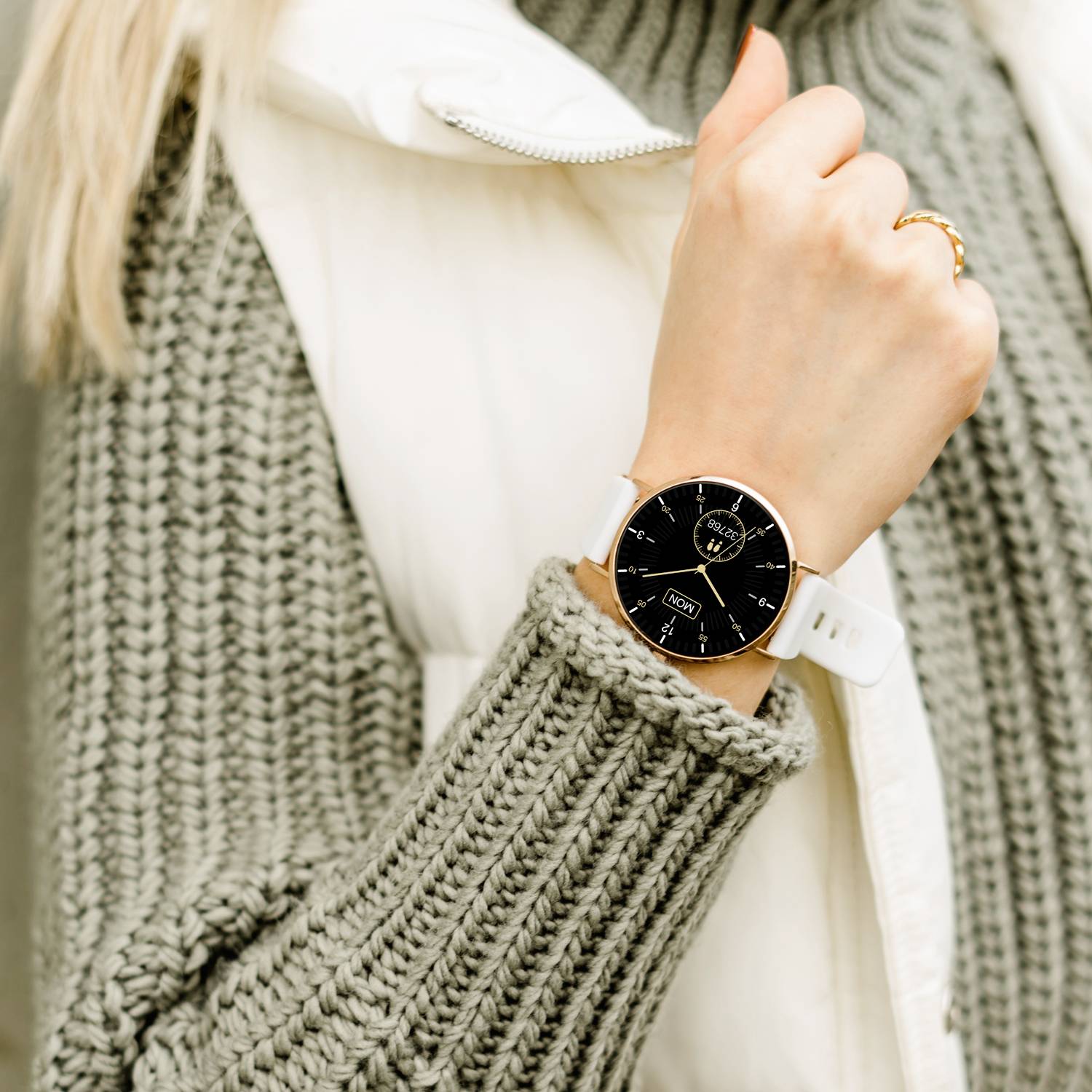 Person mit gestricktem Pullover und weißer Weste zeigt Armbanduhr mit schwarzem Zifferblatt und weißem Armband.