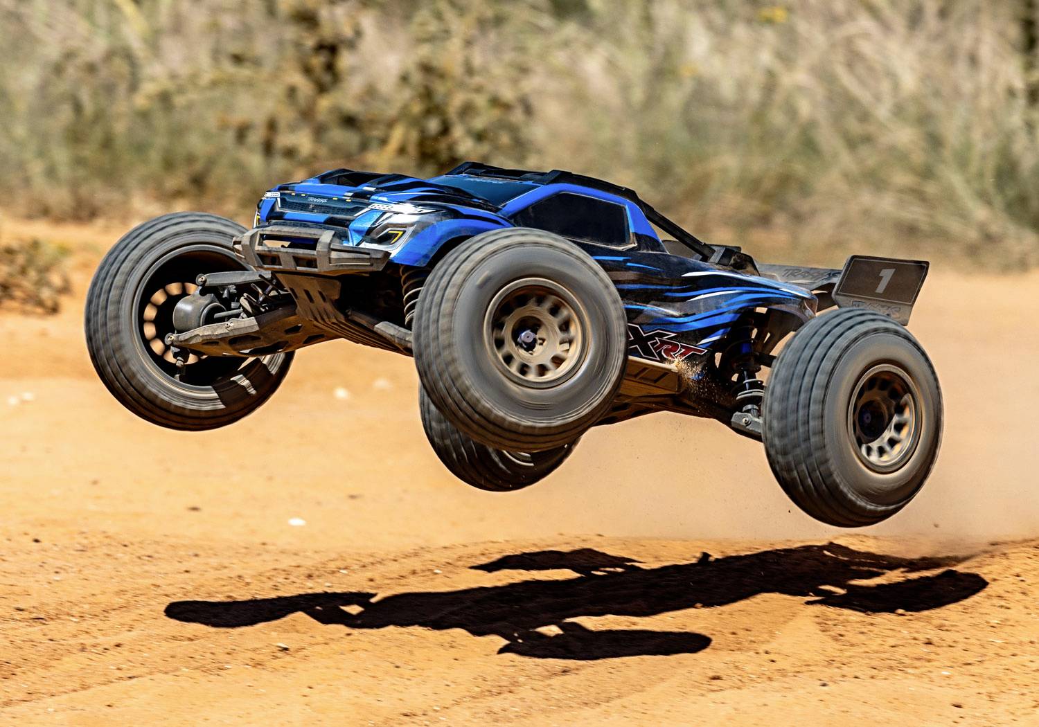 Ein blauer ferngesteuerter Truck springt dynamisch über eine sandige Piste, Staub wirbelt auf. Hintergrund unscharf mit Pflanzen.