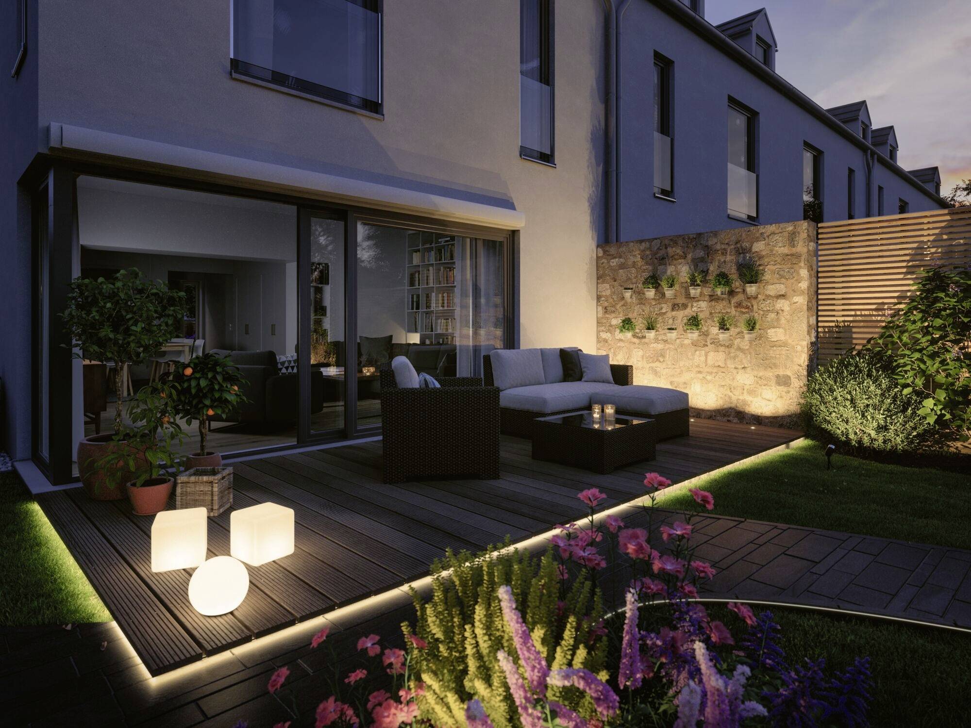 Beleuchtete Terrasse bei Dämmerung, mit modernen Möbeln, Pflanzen und dekorativen Lampen, angrenzend an ein Wohnhaus.